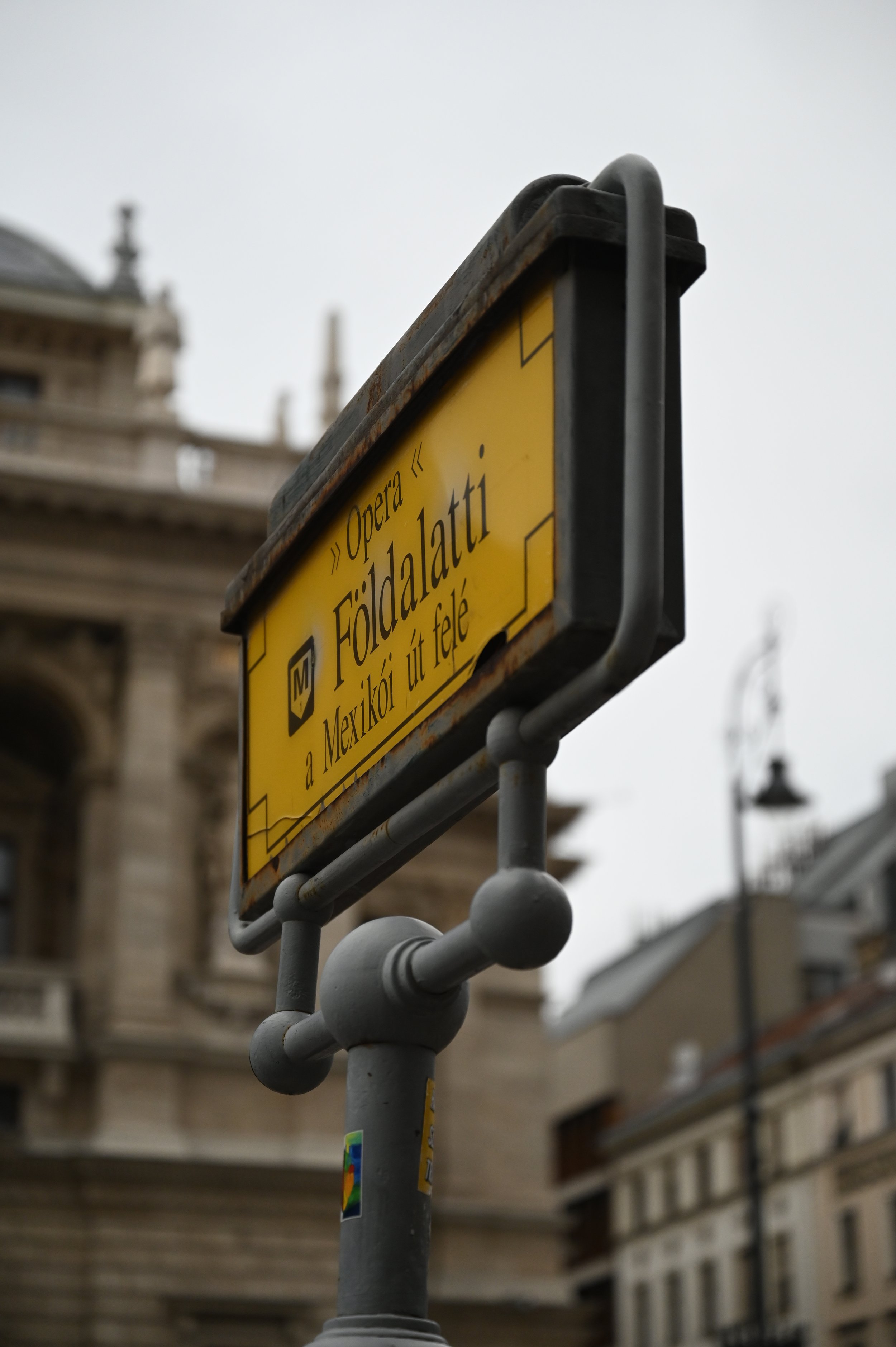 A yellow metro bus stop sign with black text and border, mounted on a gray pole with decorative elements, in an urban environment with historic buildings in the background.