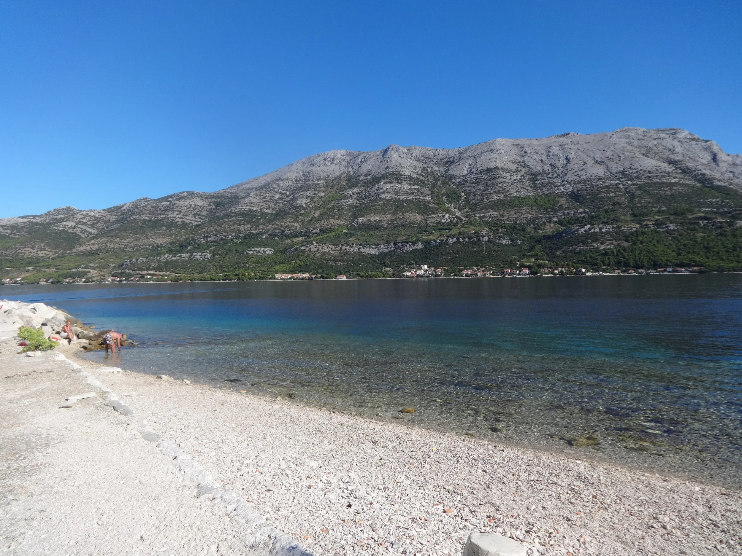 A beach with white pebbles near a body of water, with mountains in the background and a clear blue sky.