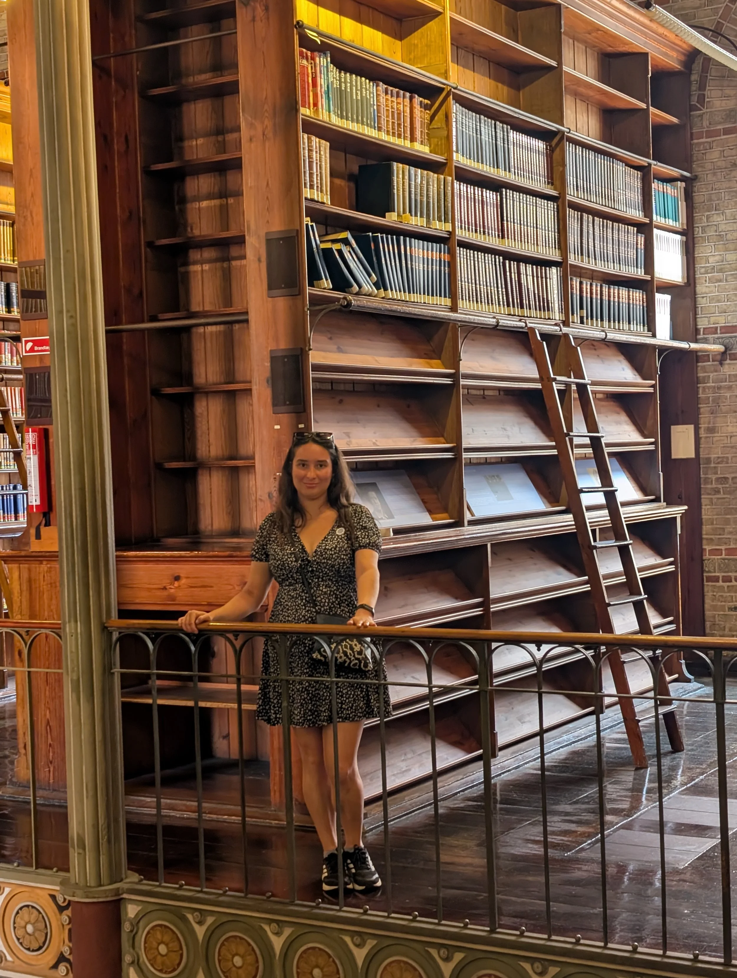 A woman standing on a balcony in front of a large wooden bookshelf inside a library. The bookshelf is mostly empty, with a few books on the upper shelves. The woman is wearing a black and white patterned dress and sneakers, and has long, dark hair.