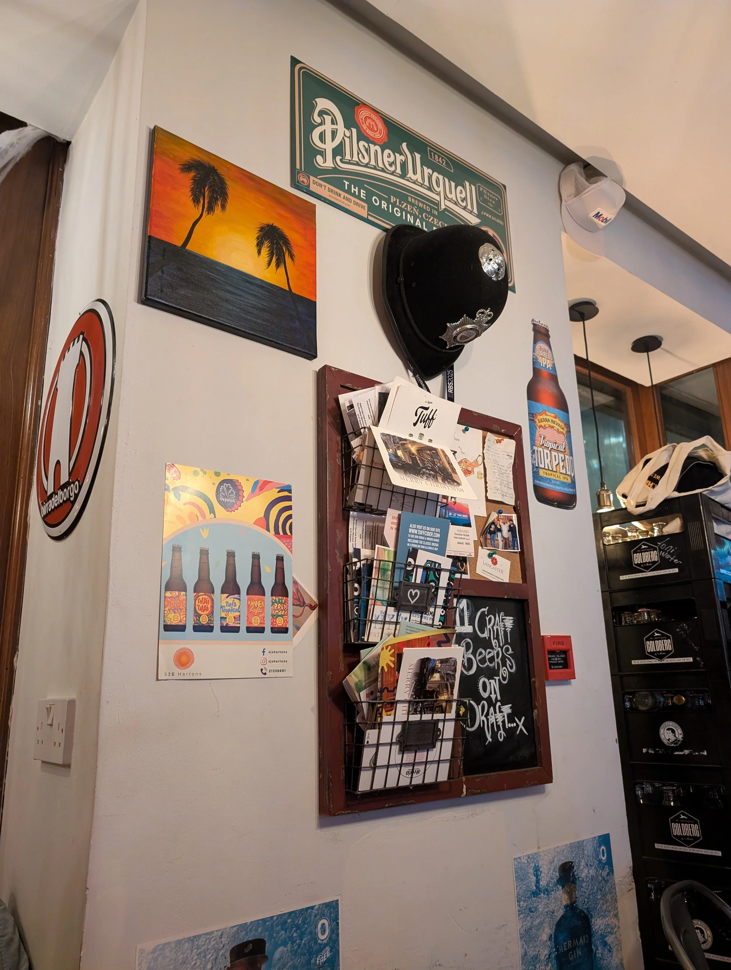 Wall decorated with various posters including a Pilsner Urquell sign, a sunset with palm trees, a poster of beer bottles, and a poster advertising craft beers on draft, with a nearby menu board and a brass bell.
