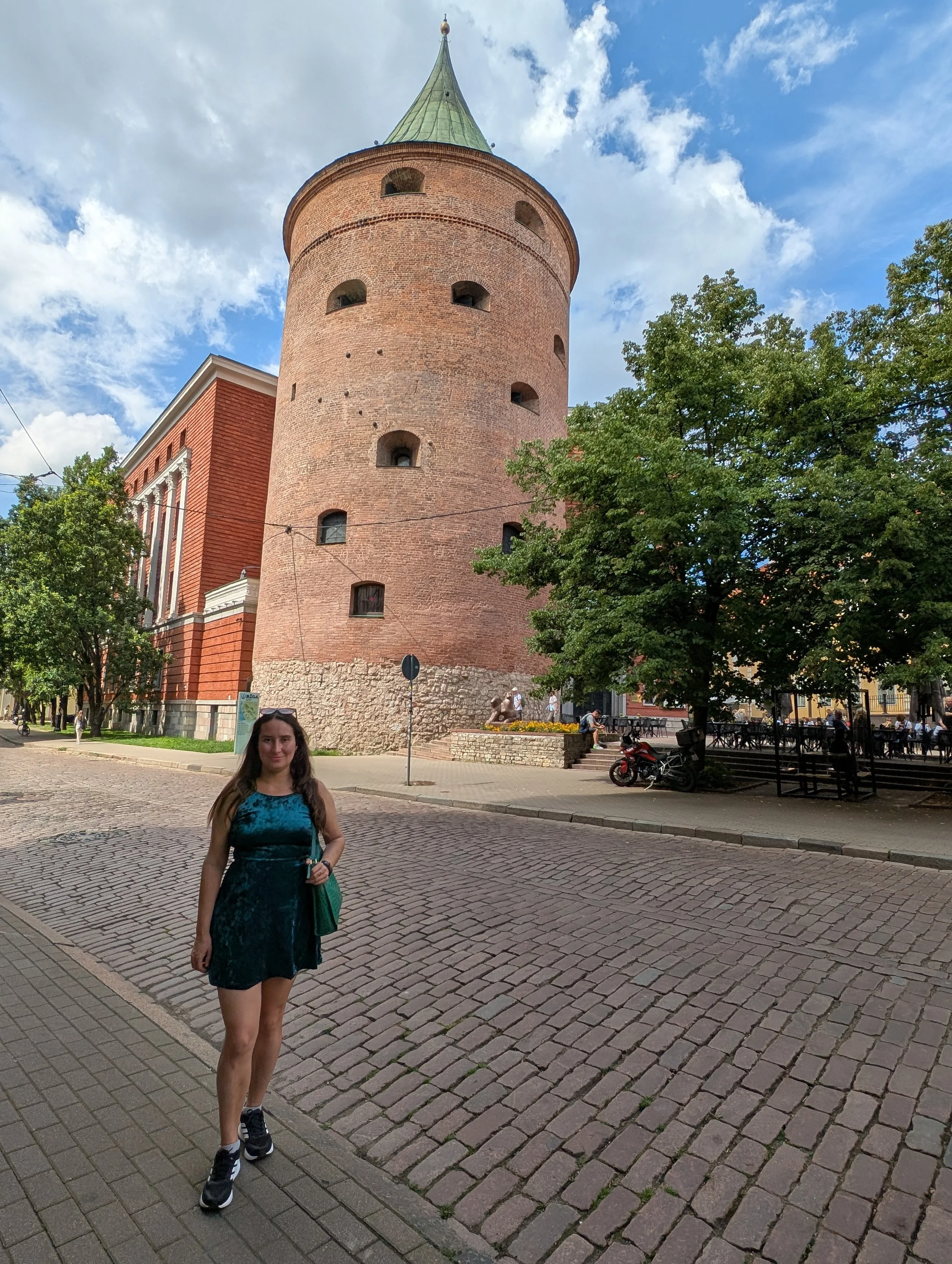 A woman wearing a denim dress and sneakers, holding a green bag, standing on a cobblestone street in front of a historic brick tower with small arched windows and a conical roof. There are trees and a small outdoor seating area with benches and table