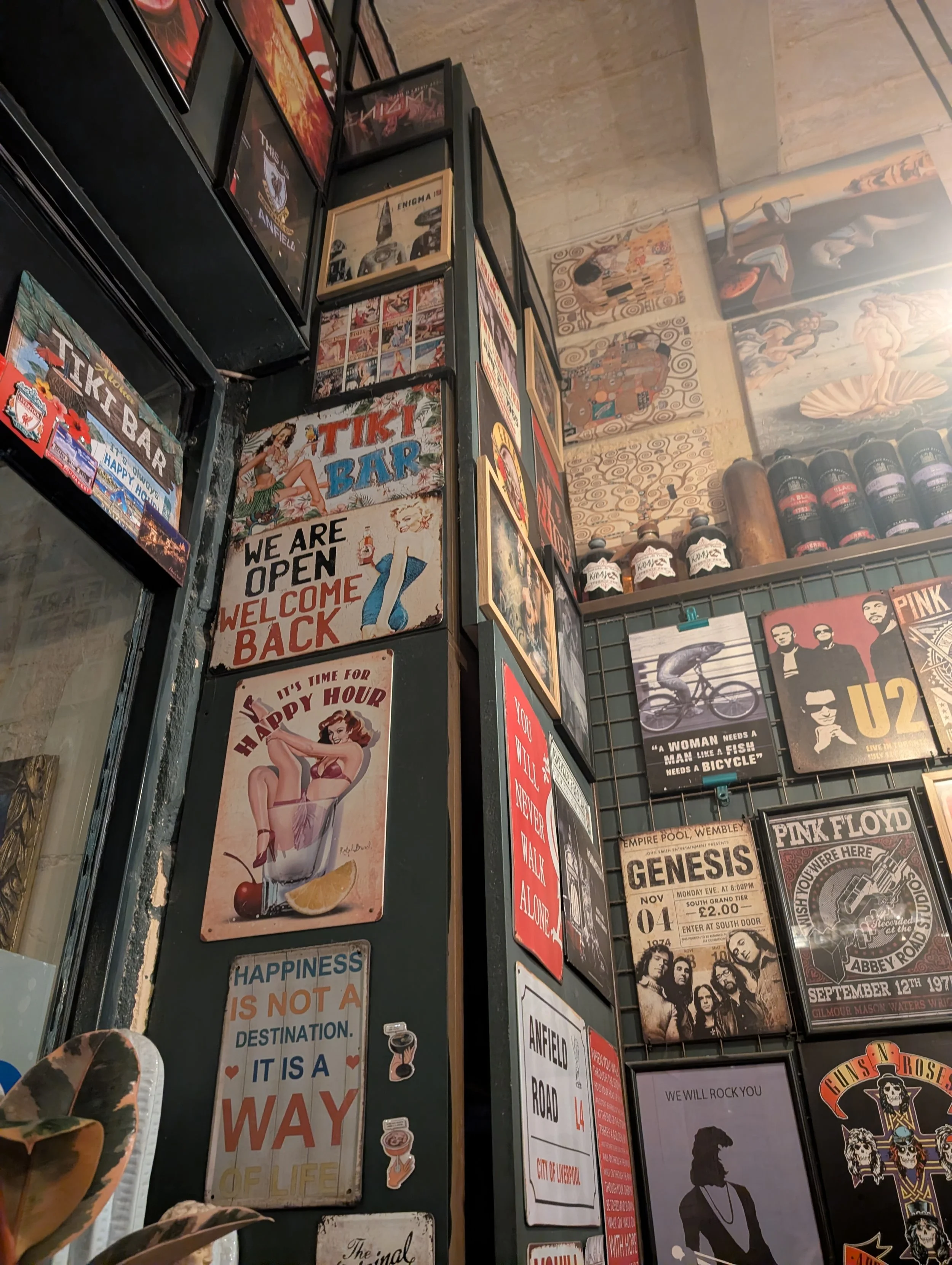 A wall display with various vintage-style posters and signs, including a 'Tiki Bar' sign, an 'We Are Open' welcome sign, and posters of bands like U2 and Pink Floyd, with some paintings and decorative art on the upper part of the wall.