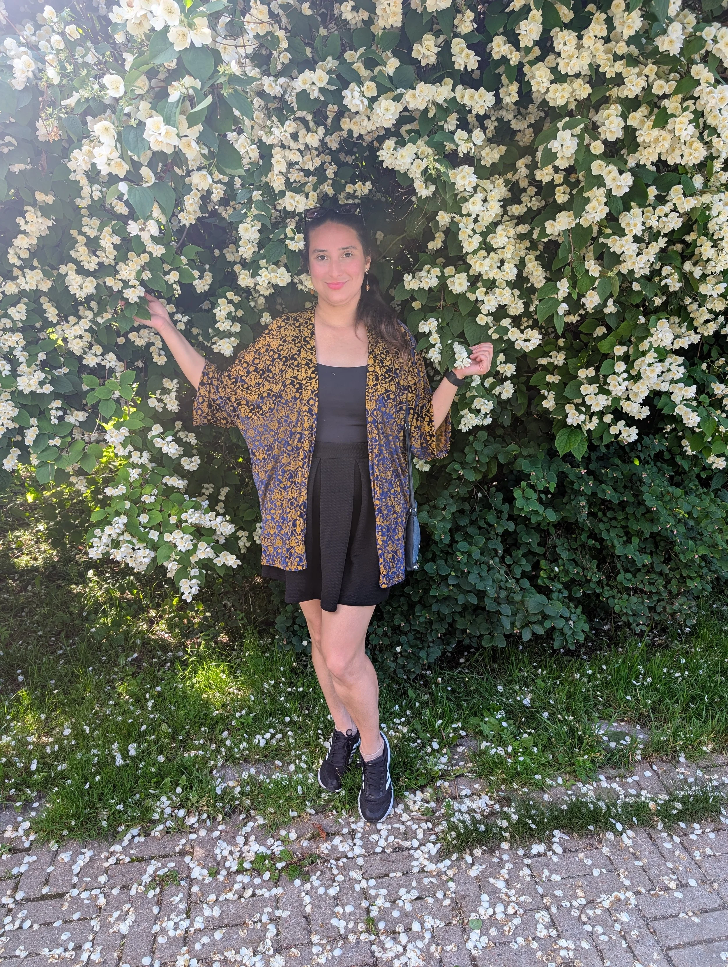 A woman standing outdoors, smiling, with her arms raised, surrounded by white flowers on a bush. She is wearing a black top, shorts, a patterned jacket, and sneakers, with sunglasses on her head and earrings.
