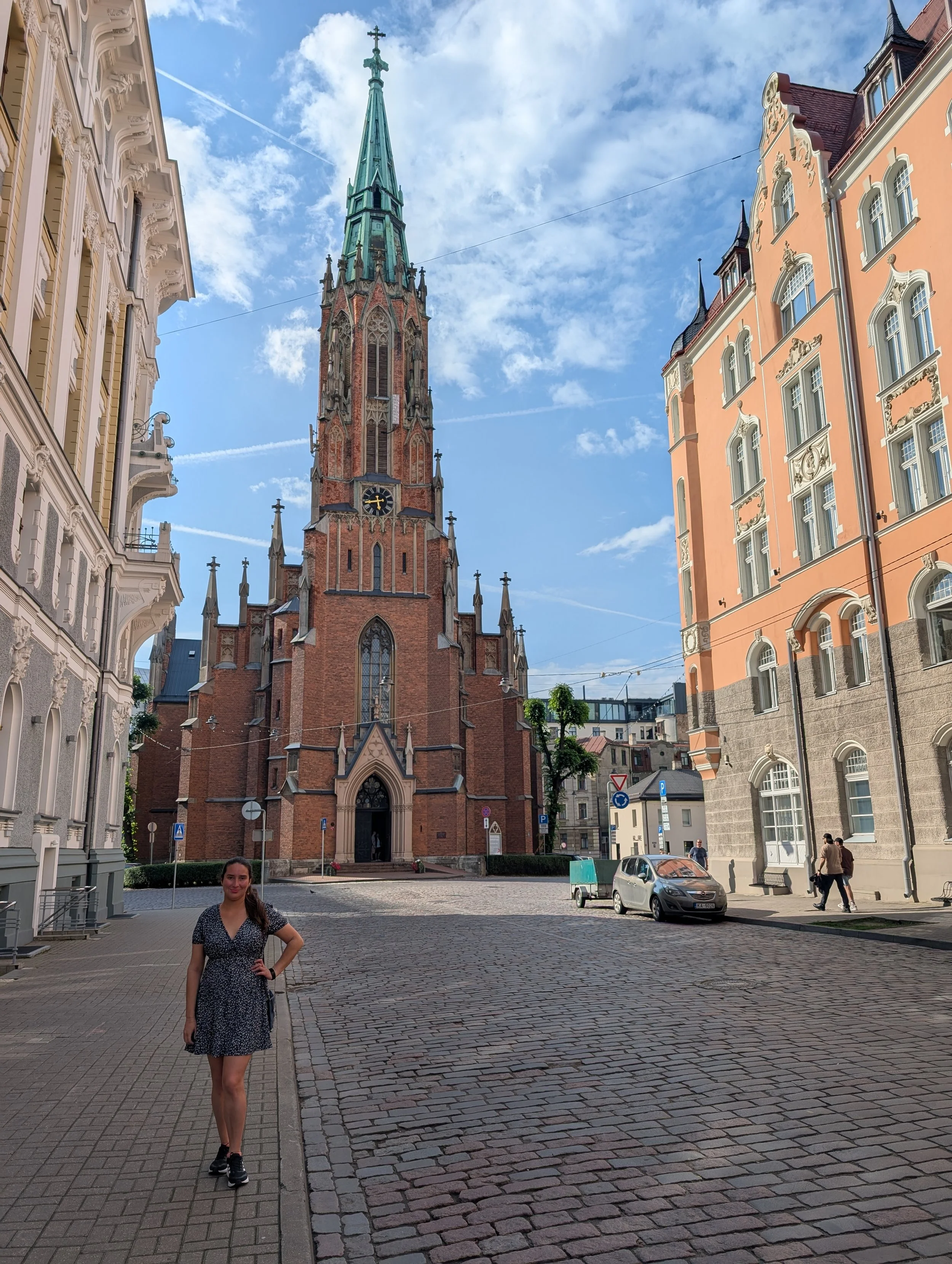 A woman in a black and white polka dot dress standing on a cobblestone street near historic buildings, with a church steeple in the background under a partly cloudy sky.