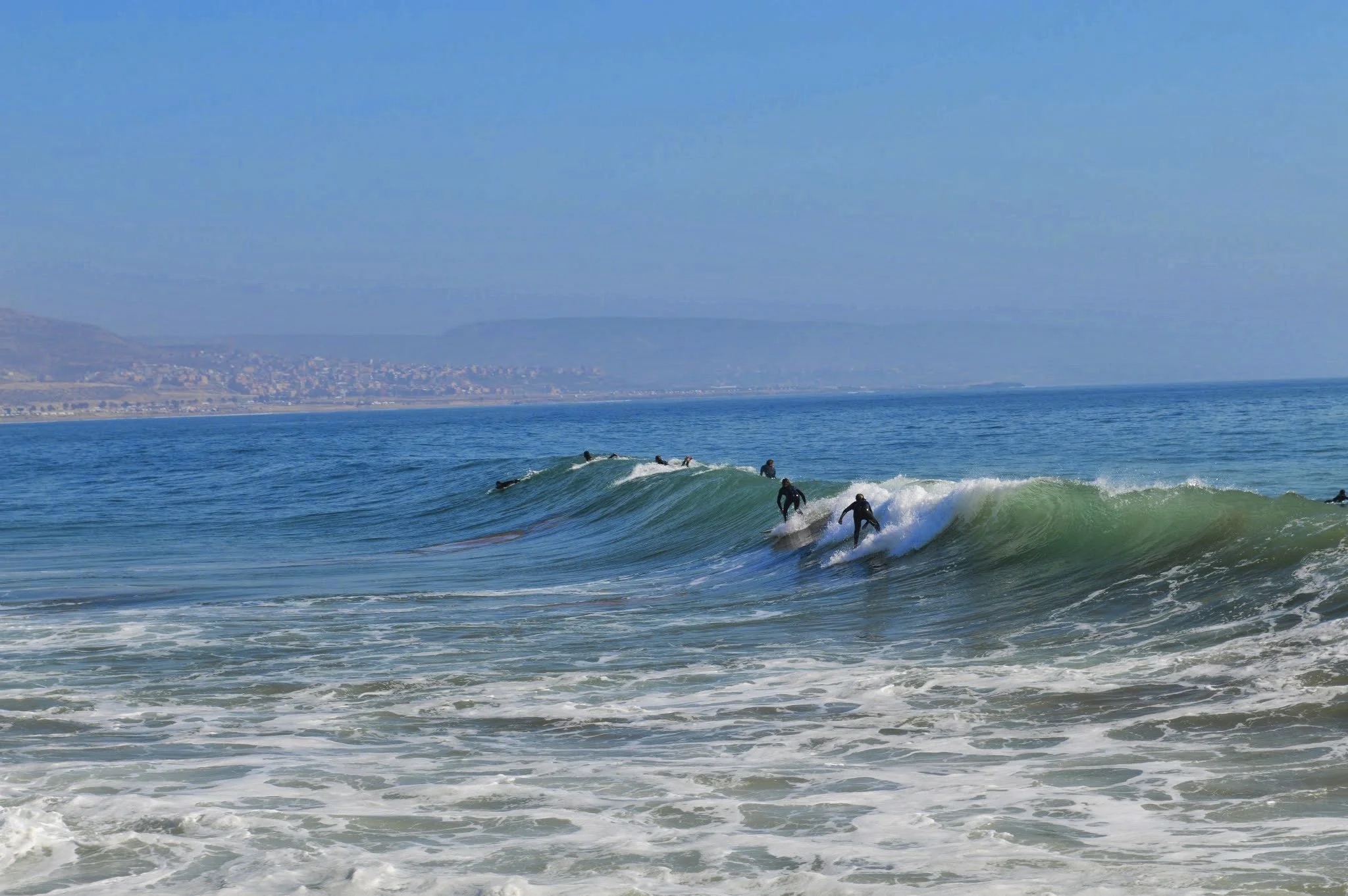 Surfers riding in the ocean waves near a coastline with a distant view of a town and mountains under a clear blue sky.