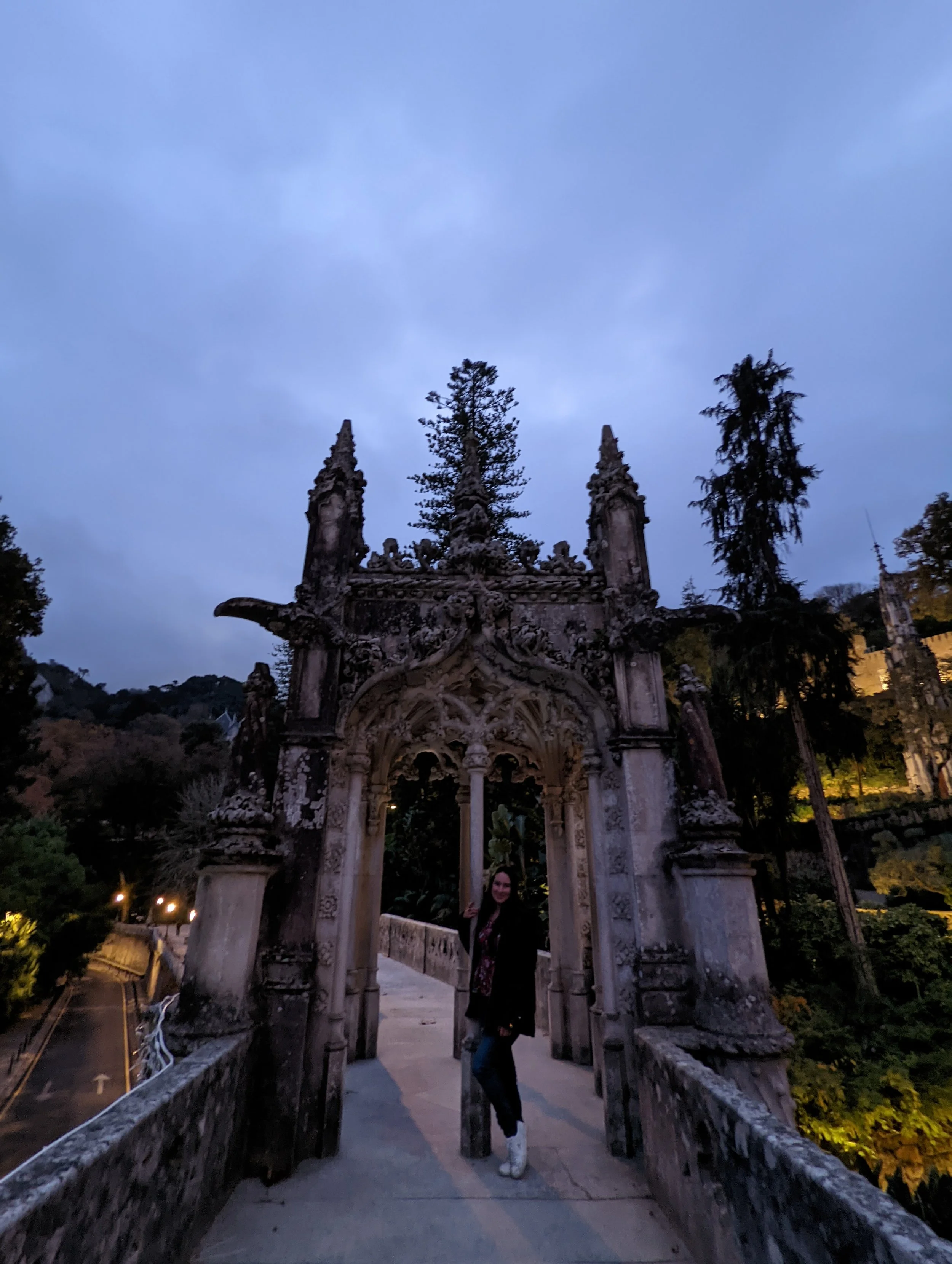 A woman standing under a historic ornate stone archway at dusk, with trees and a cloudy sky in the background.