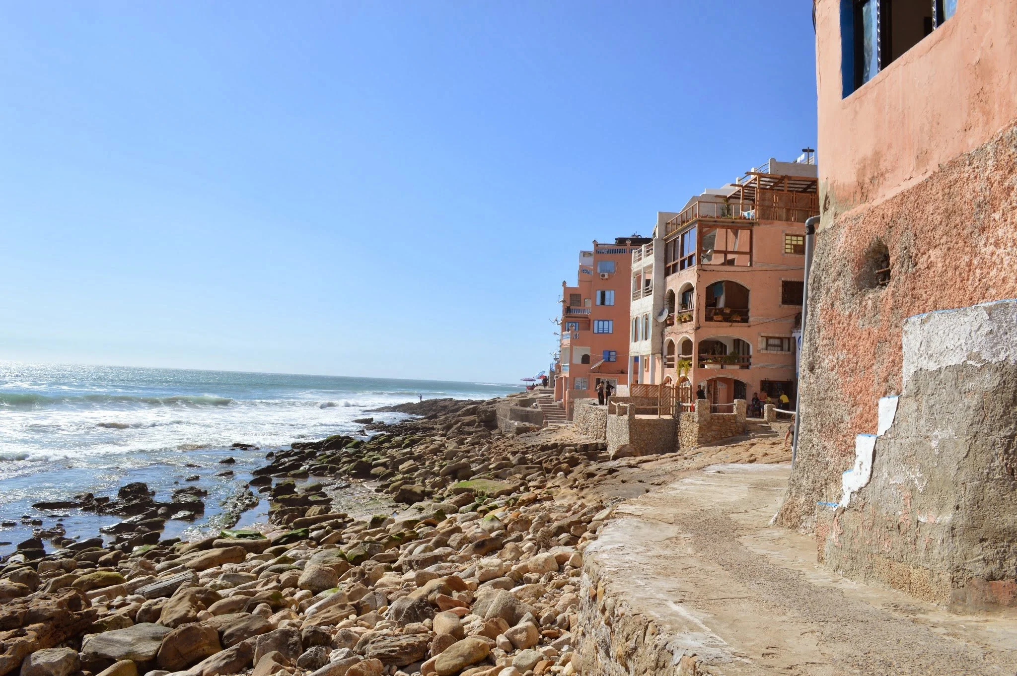 Rocky beach with a row of pastel-colored buildings along the shoreline, overlooking the ocean under a clear blue sky.