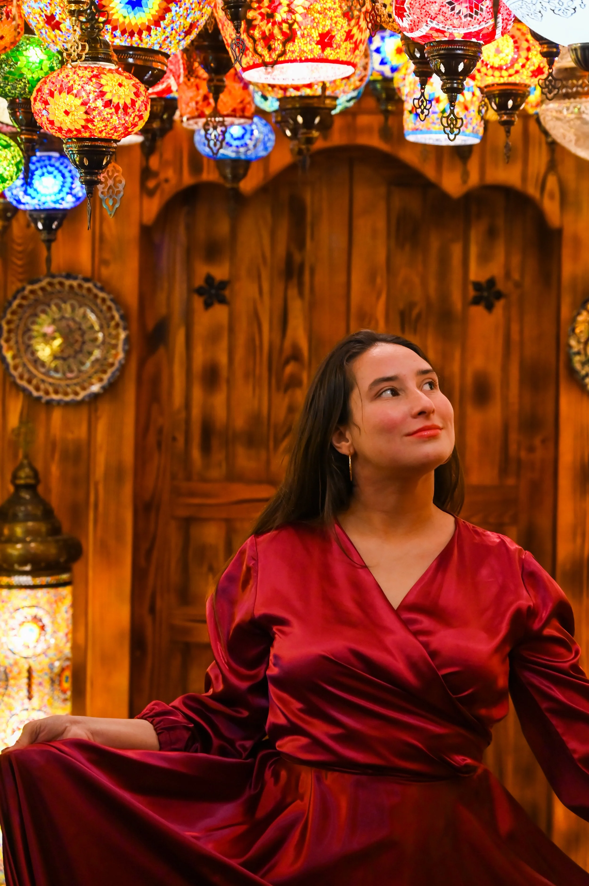 A woman in a red satin dress sitting in a room with wooden walls and colorful Turkish mosaic lamps hanging from the ceiling.