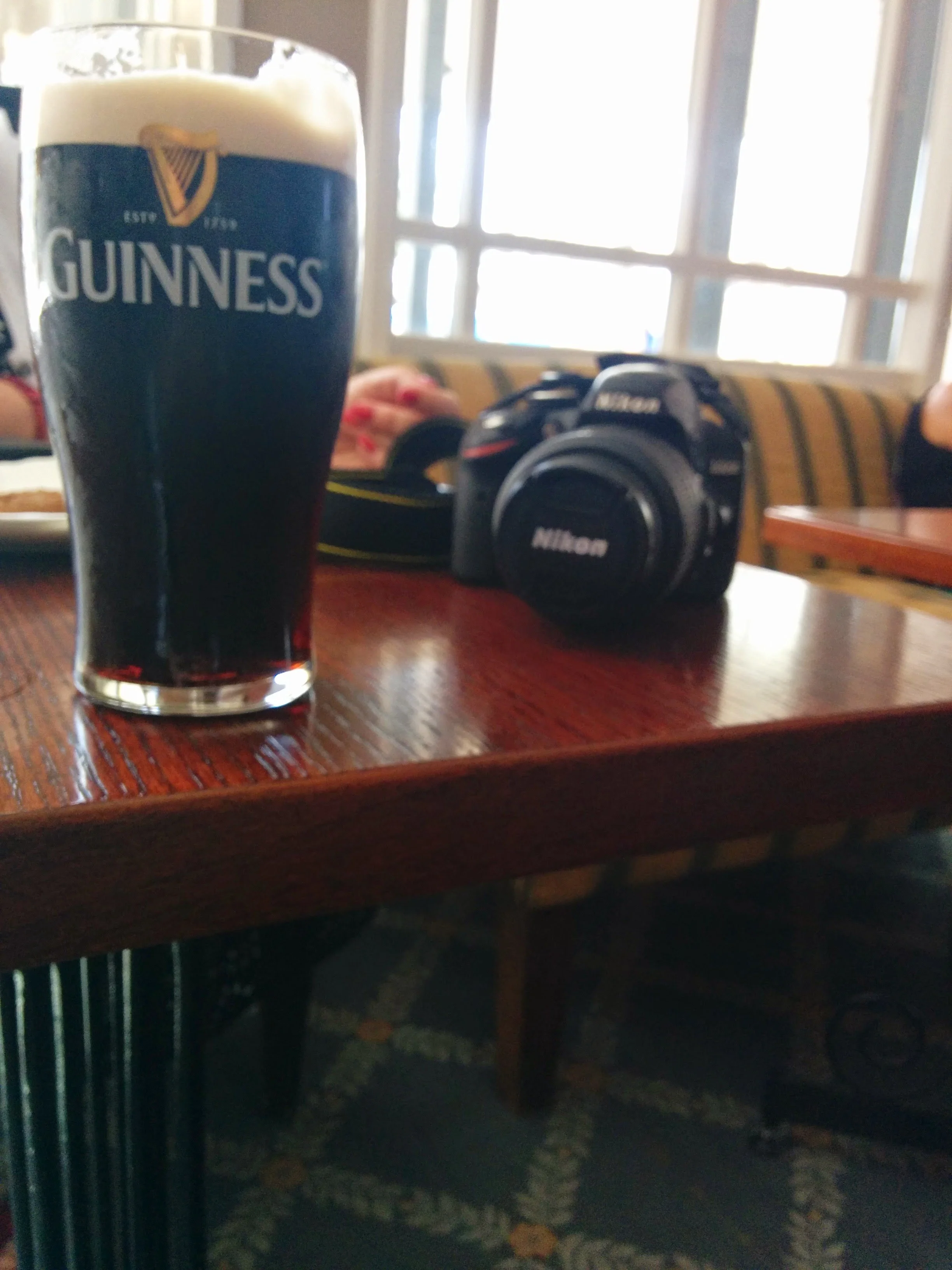 A pint glass of Guinness beer on a table, with a camera and a person's hands in the background, near a window with sunlight.