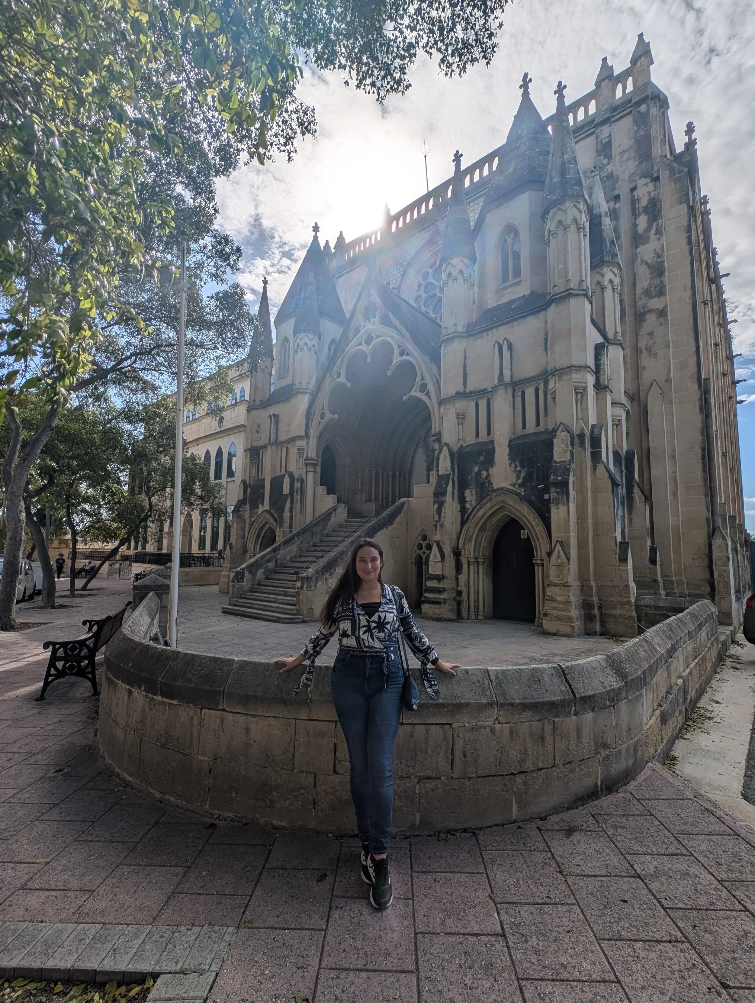 A woman with long dark hair wearing a patterned jacket and jeans is standing in front of a historic Gothic-style church with stone steps and a circular stone barrier. The church has pointed arches, stained glass windows, and spires. The scene is outd