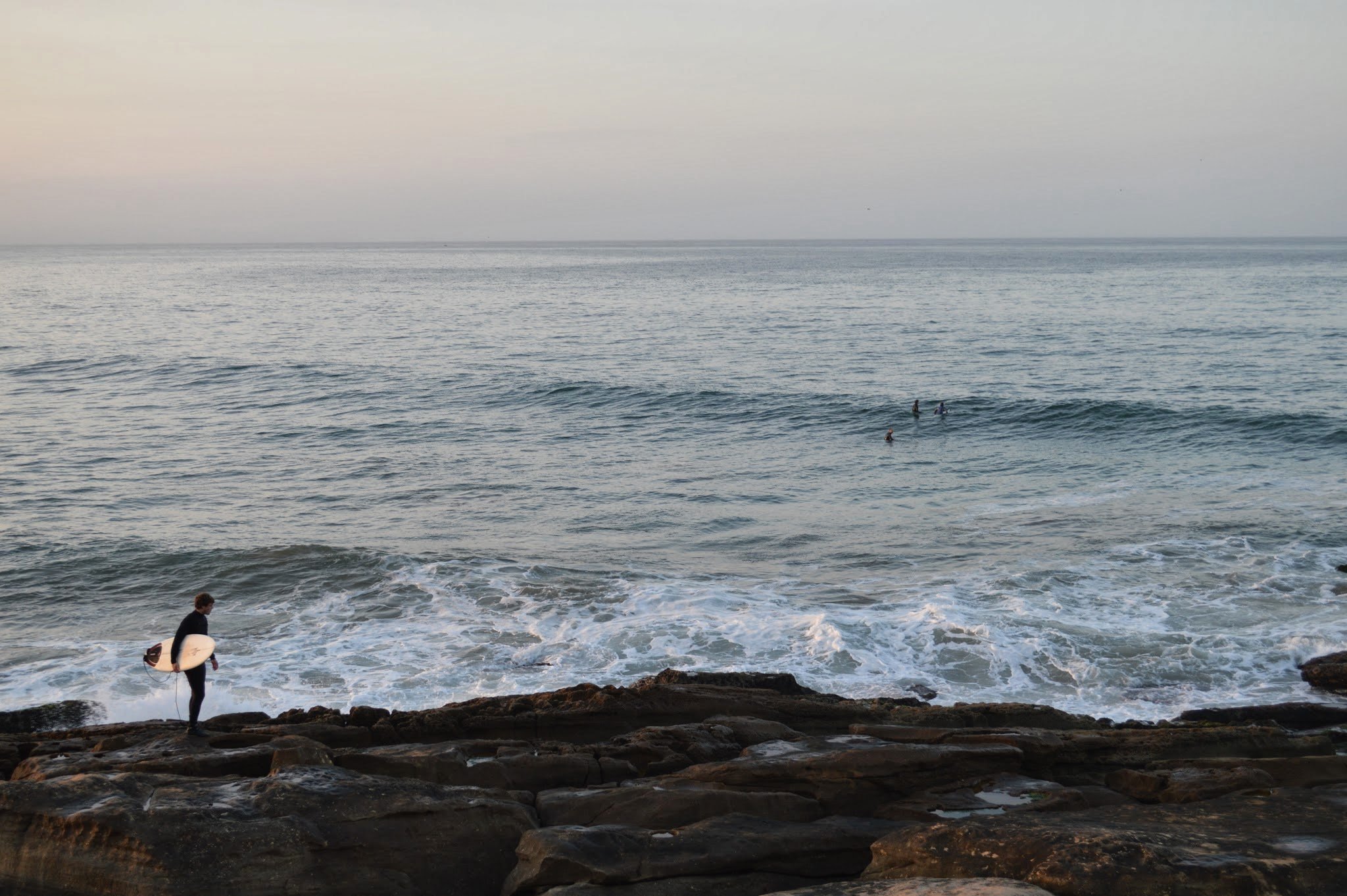 Person holding a surfboard at the rocky shoreline with people in the ocean and an overcast sky in the background.