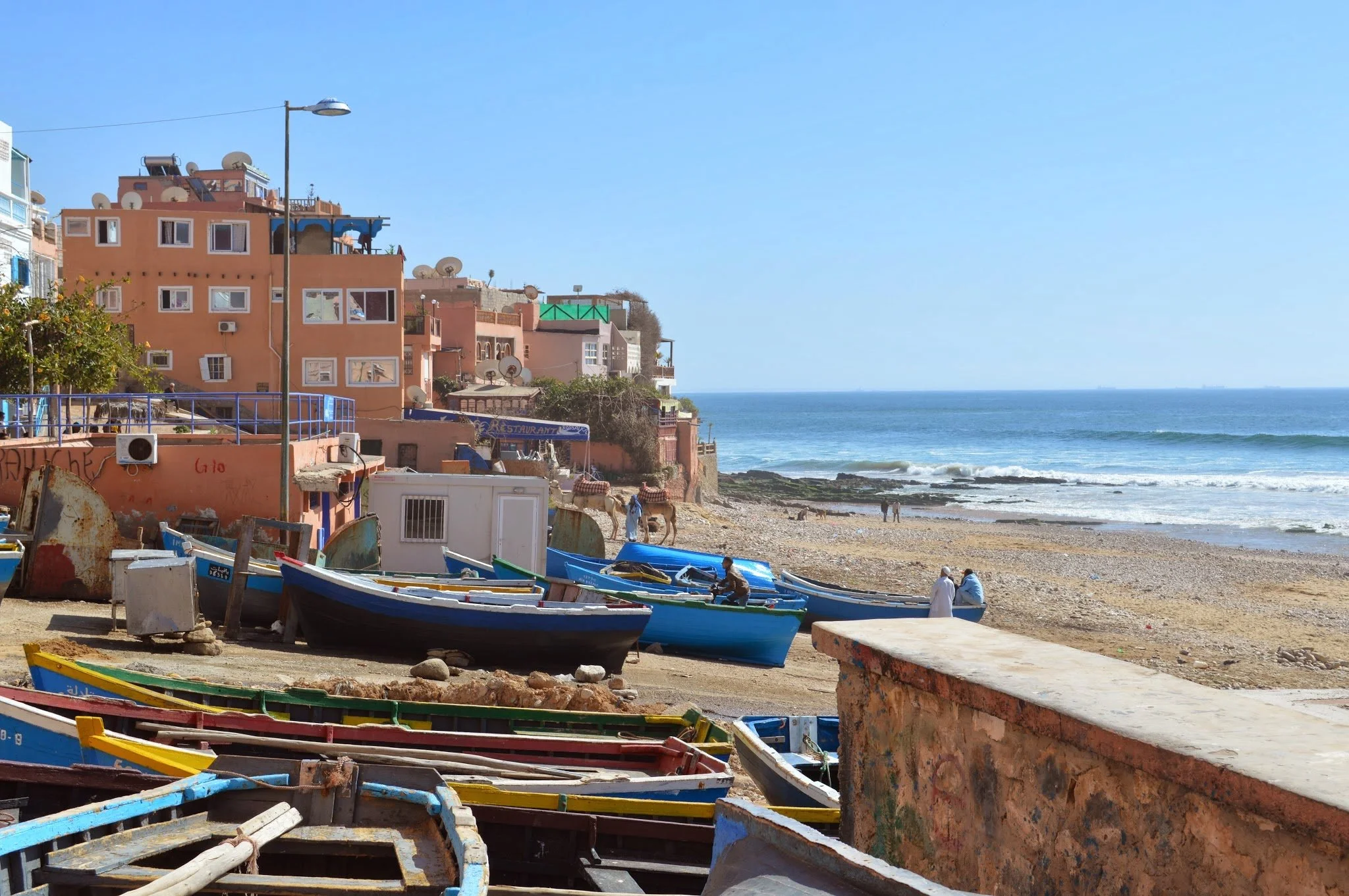 Colorful boats on a beach with pink and orange buildings on a hill in the background and the ocean in the distance.