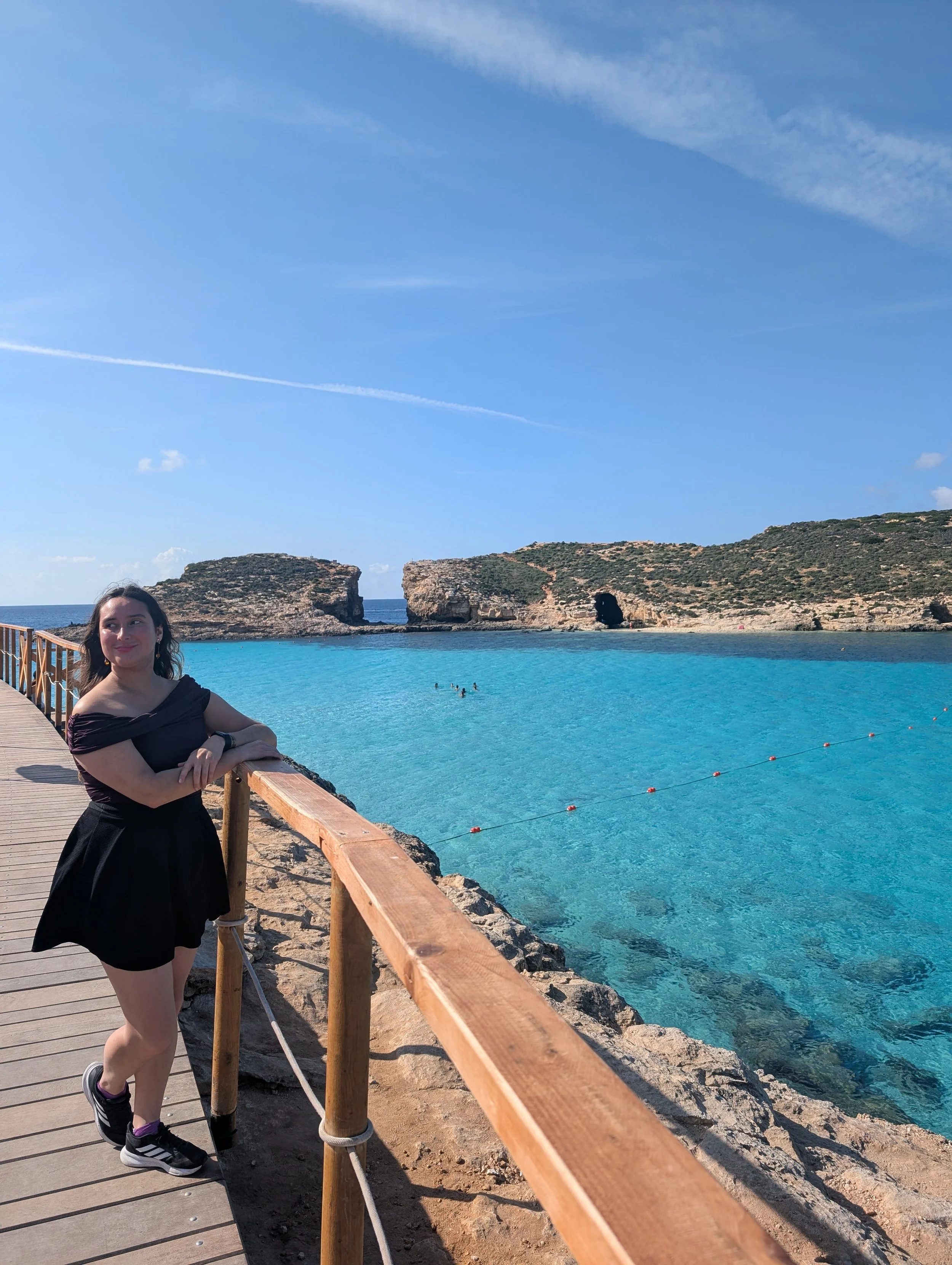 A young woman standing on a wooden walkway by a turquoise beach, with rocky cliffs and blue sky in the background.