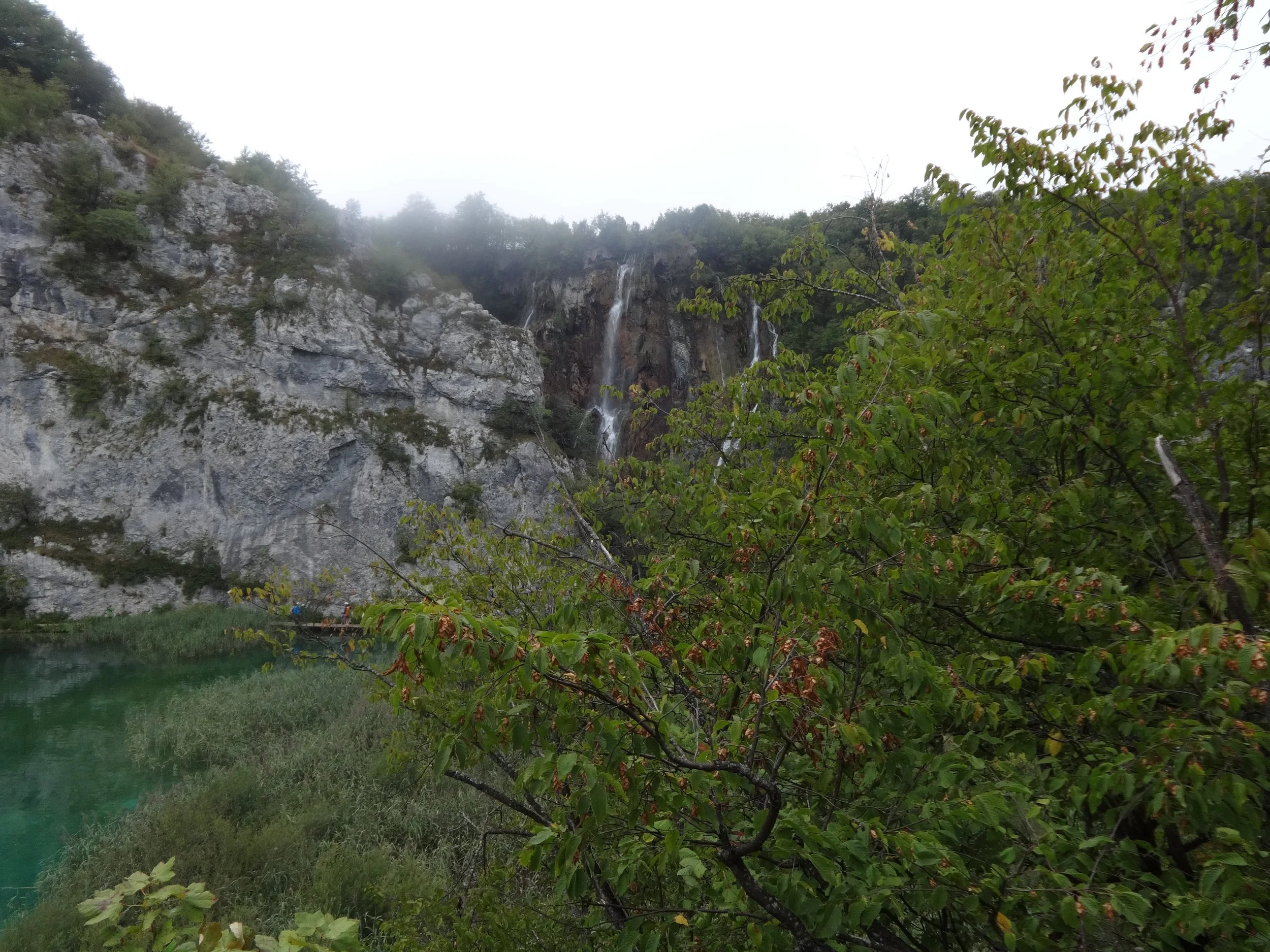 A scenic view of a mountain with waterfalls, trees, and green foliage near a body of water, under a cloudy sky.