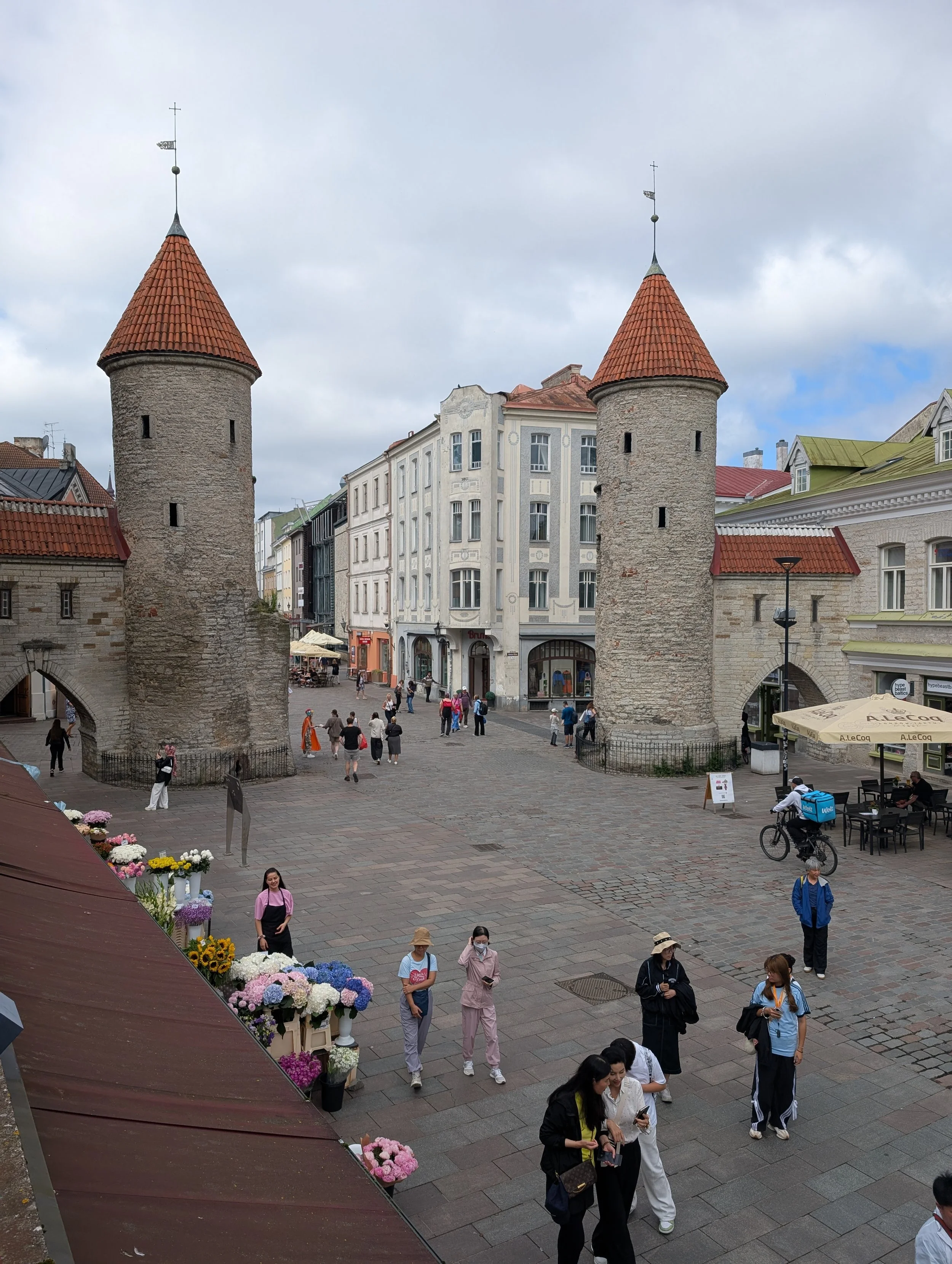 A busy plaza with people walking and shopping near historic stone towers with red tile conical roofs. The scene includes shops, outdoor seating, a bicycle courier, and flower displays.