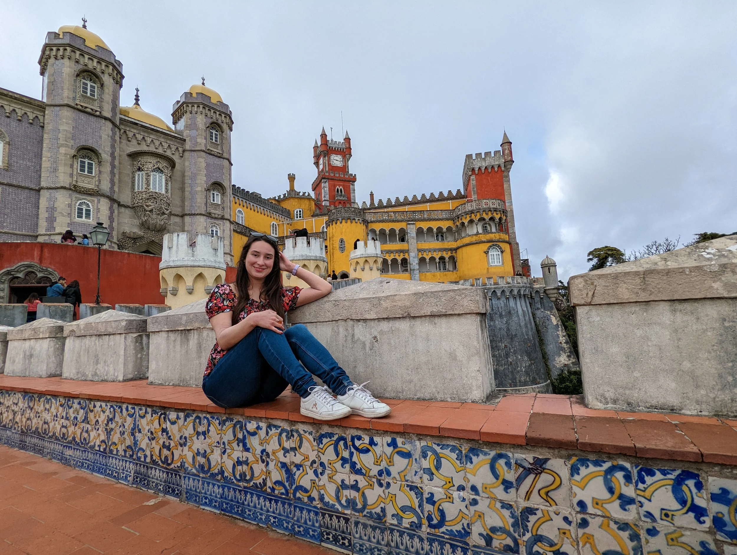 A woman sitting on a decorated ledge in front of colorful castle-like architecture with towers and turrets, including gold domes and a red clock tower.
