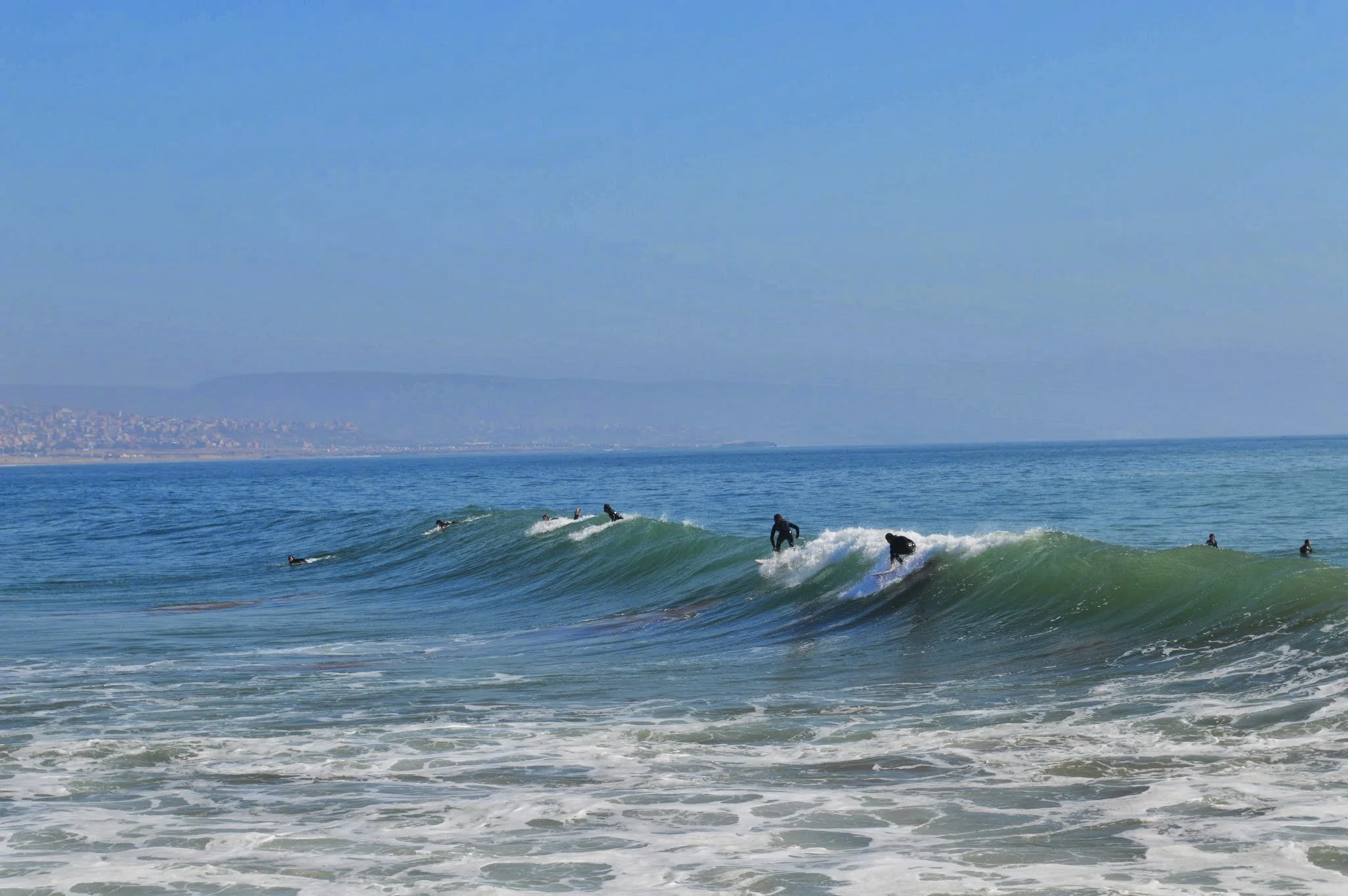 Surfers riding waves in the ocean with a distant landmass and clear blue sky in the background.