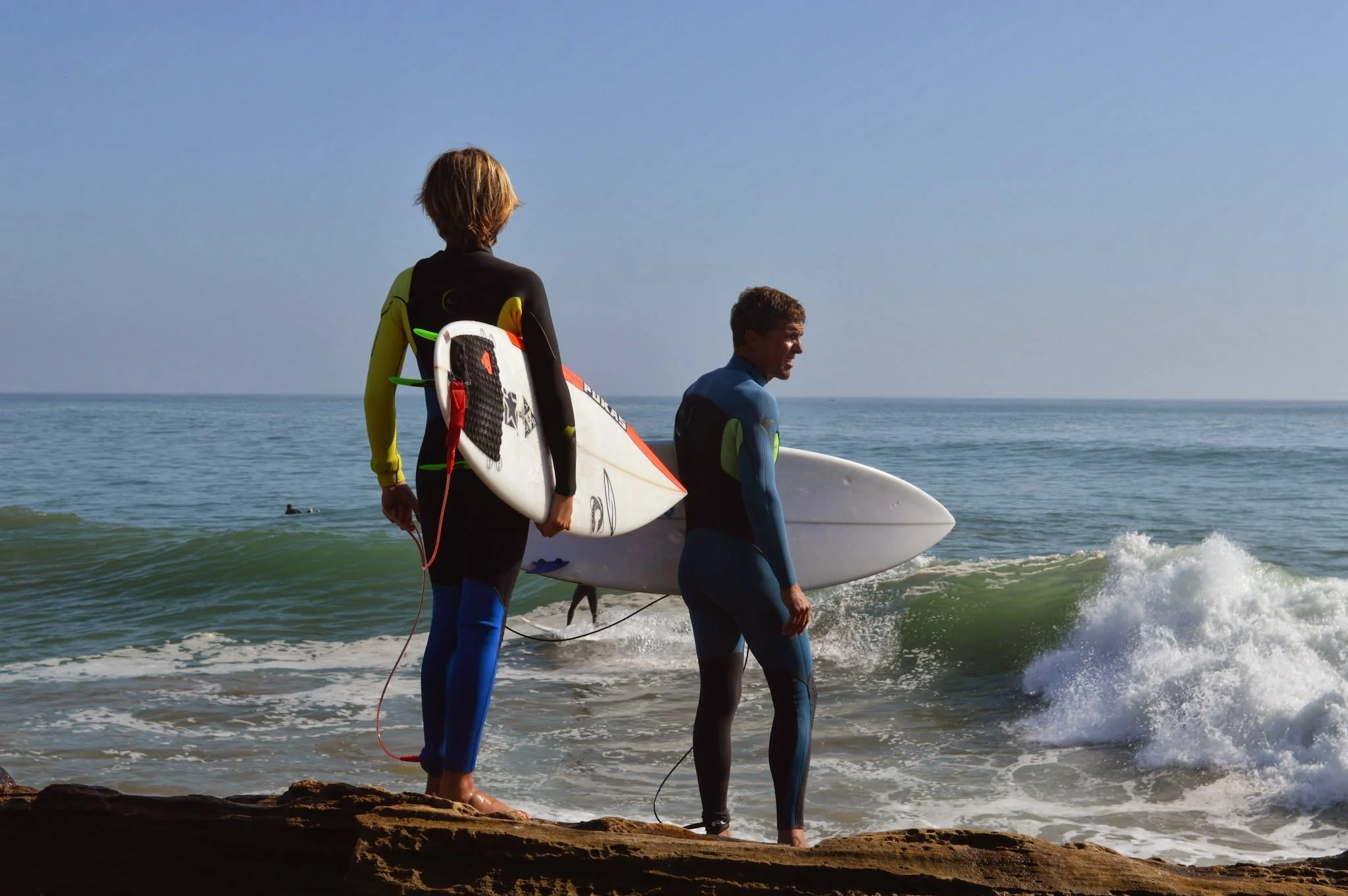 Two surfers wearing wetsuits walk along the shoreline carrying surfboards, with the ocean and clear blue sky in the background.