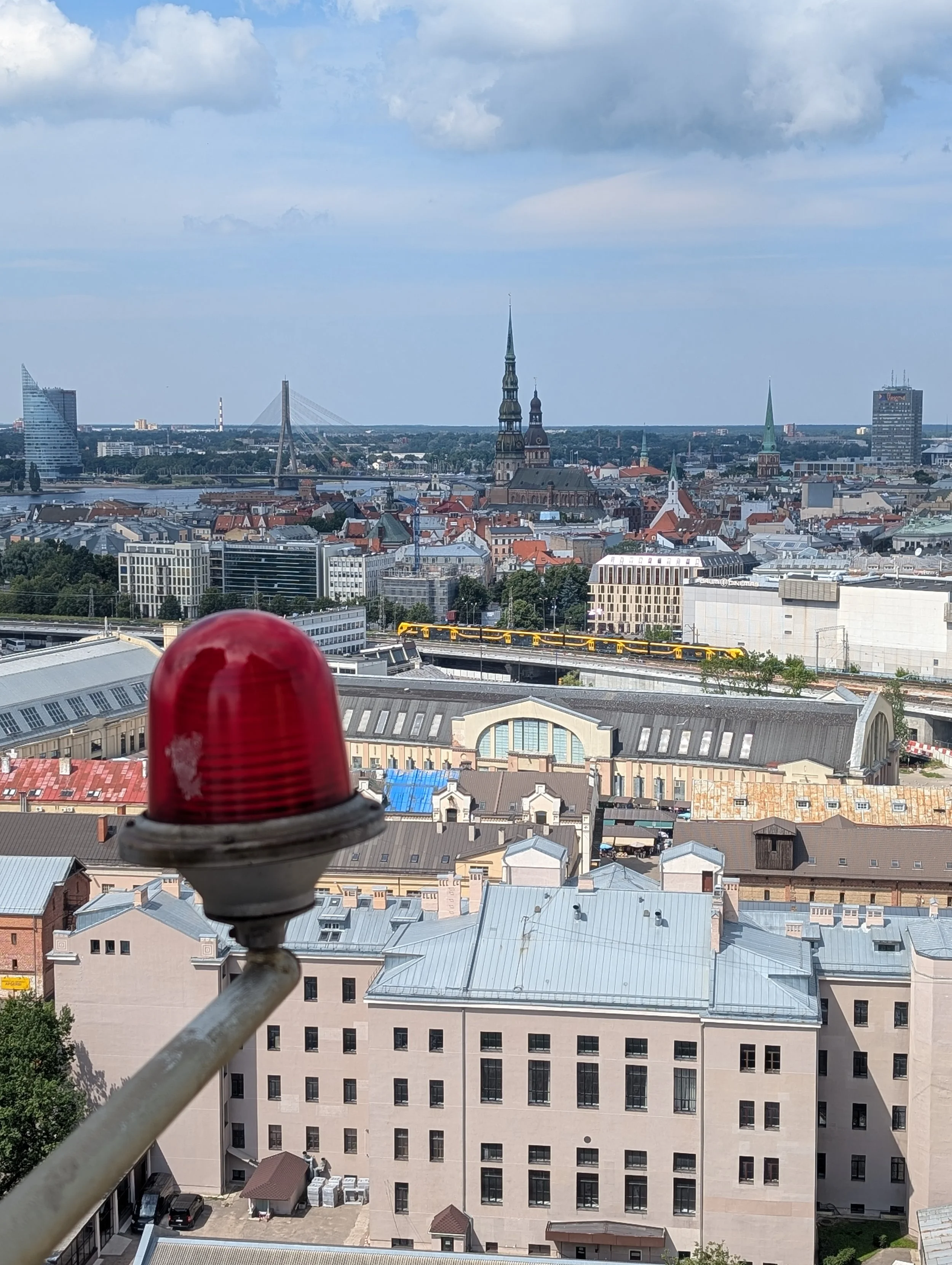 A city skyline featuring a river, historic church steeples, modern buildings, and a suspension bridge, with a red warning light in the foreground.