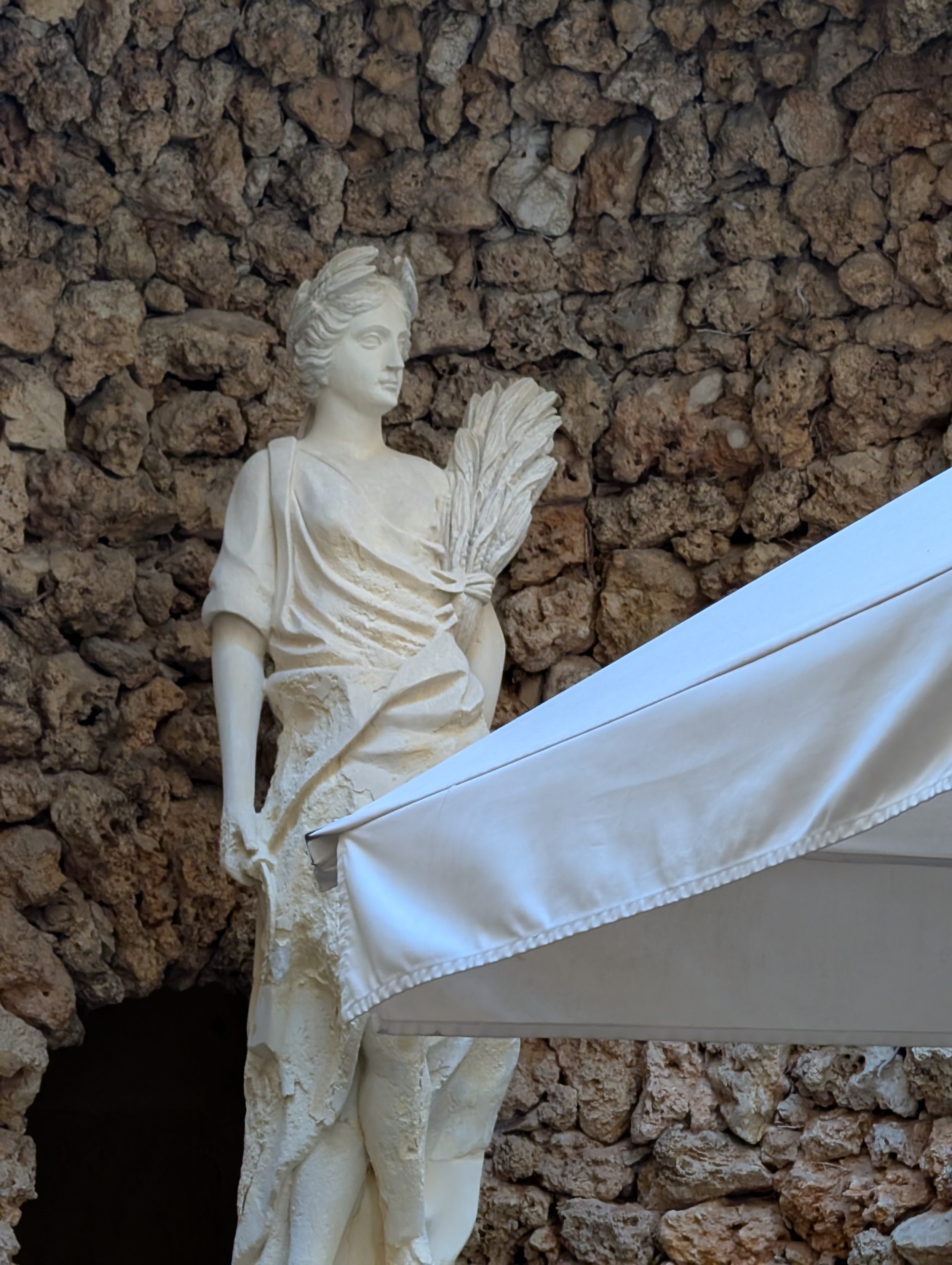 A white statue of a woman holding a bouquet of flowers, placed against a stone wall, with part of a white cloth visible at the bottom right corner.
