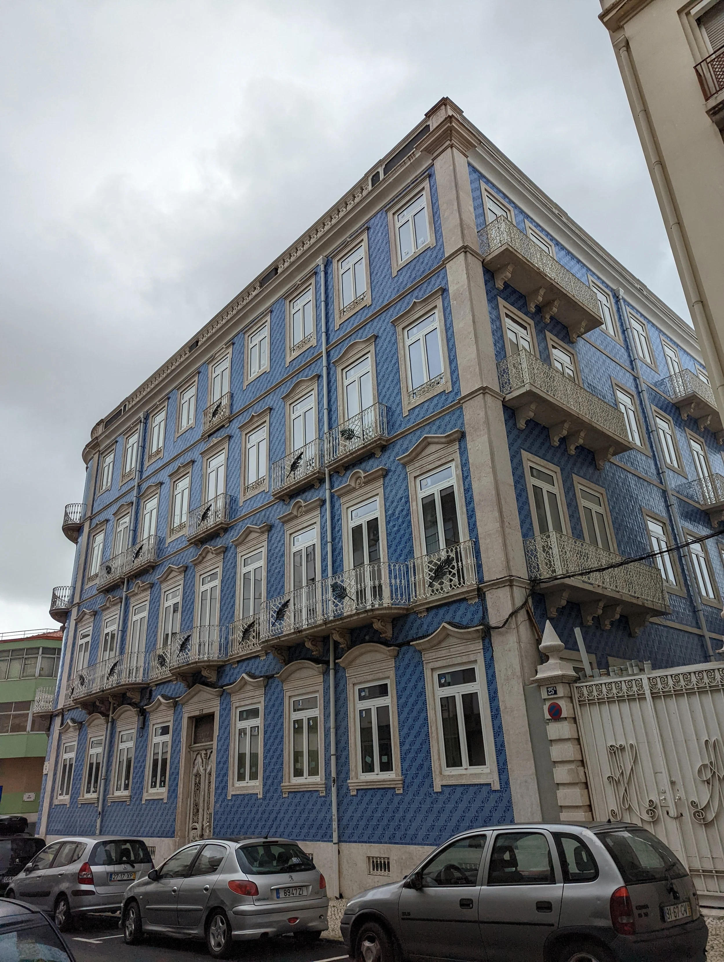 A multi-story building with a blue decorative facade, white window frames, and balconies. Several cars are parked in front of the building under a cloudy sky.