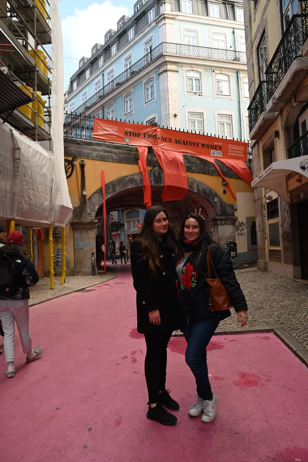 Two women standing together on a pink street in an urban area, with a bridge and buildings above. A red banner with black text about stopping violence against women is visible above them.