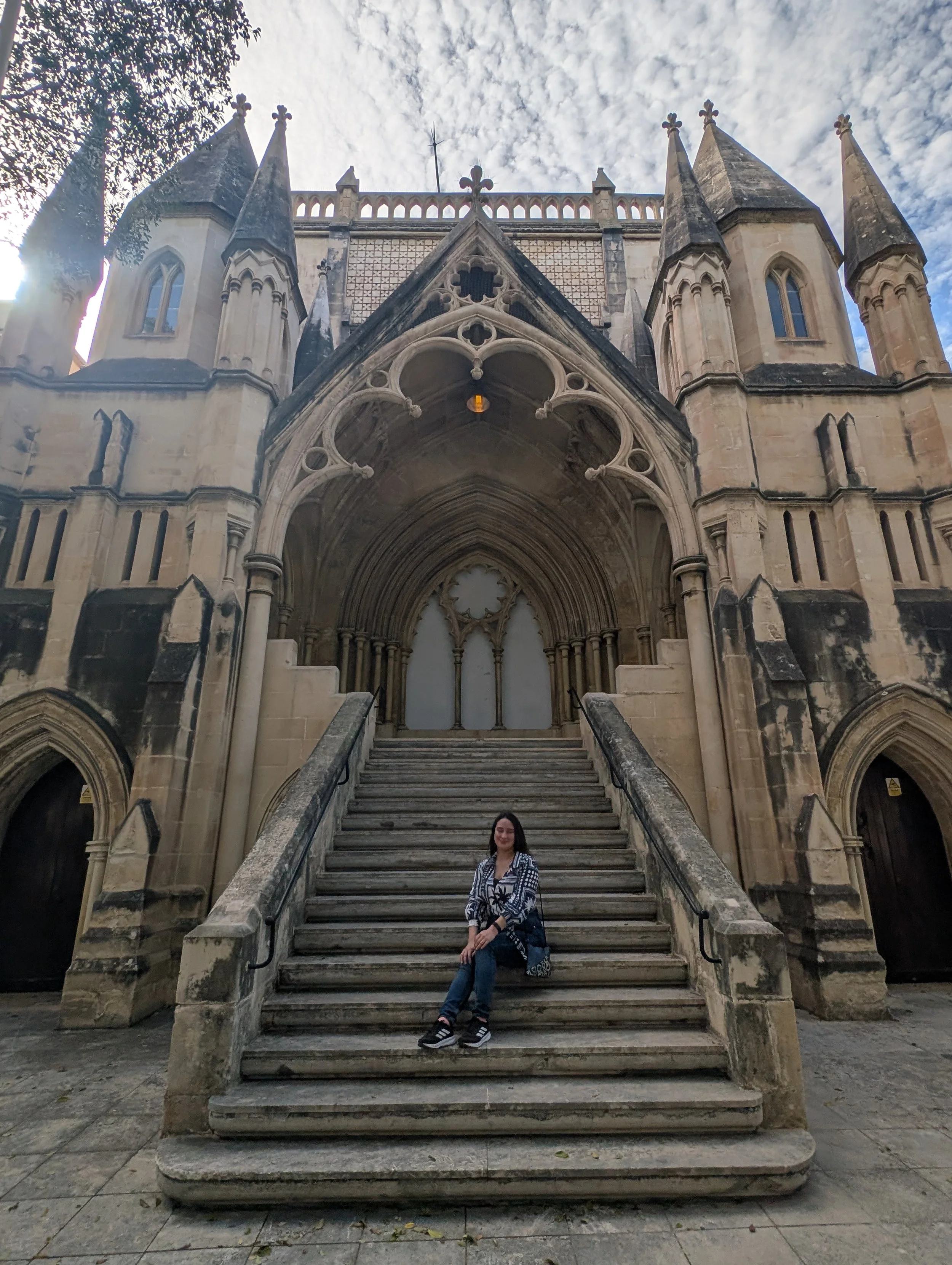 A woman with long dark hair wearing a black and white patterned blouse and jeans sitting on the steps of a historic gothic cathedral with pointed arches, spires, and intricate stone carvings, under a partly cloudy sky.