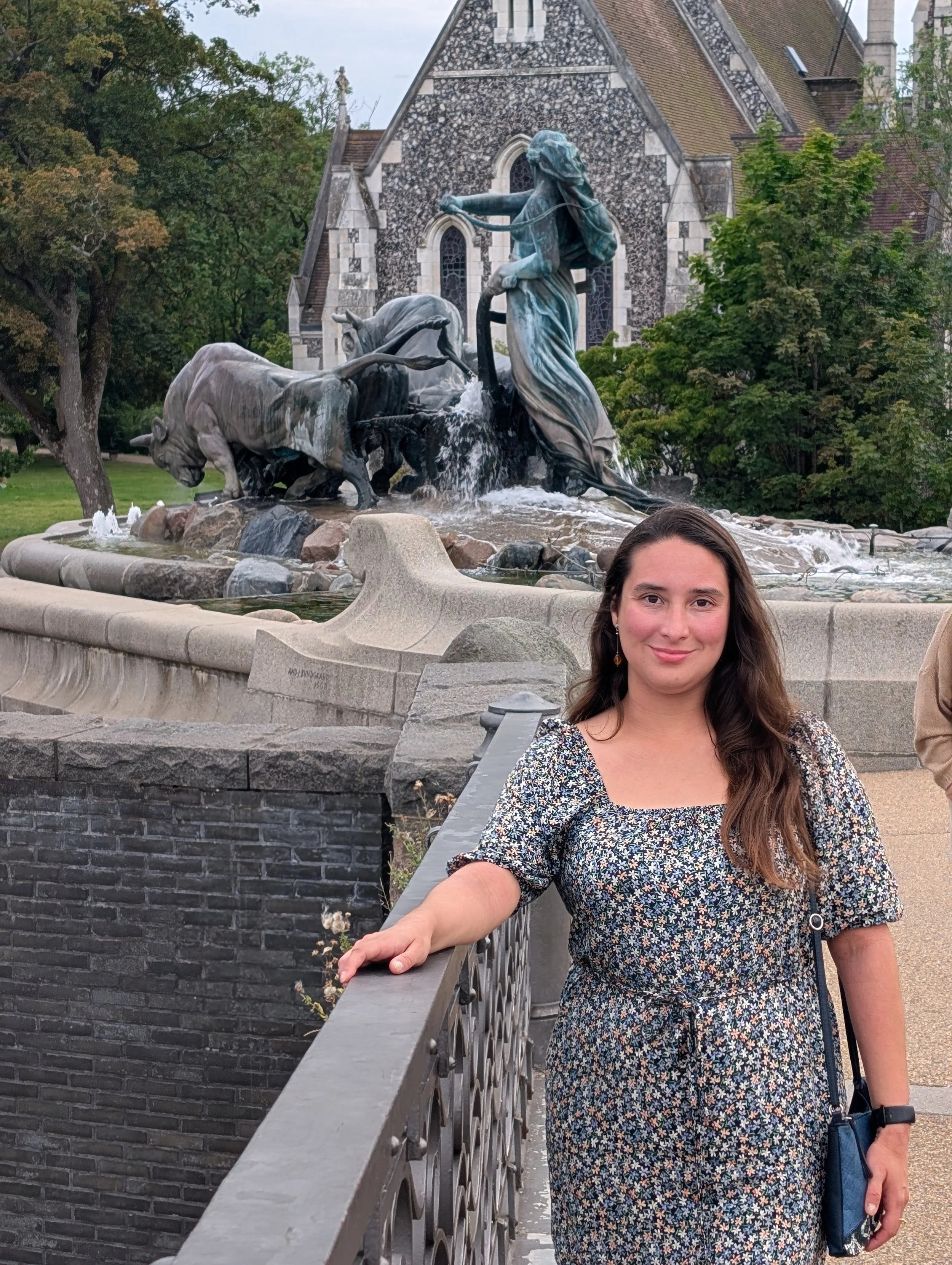 A woman with long brown hair, wearing a floral dress, leaning on a stone railing in front of a large fountain with a statue of a woman and horses, located in a park or courtyard near a stone church with stained glass windows and trees.