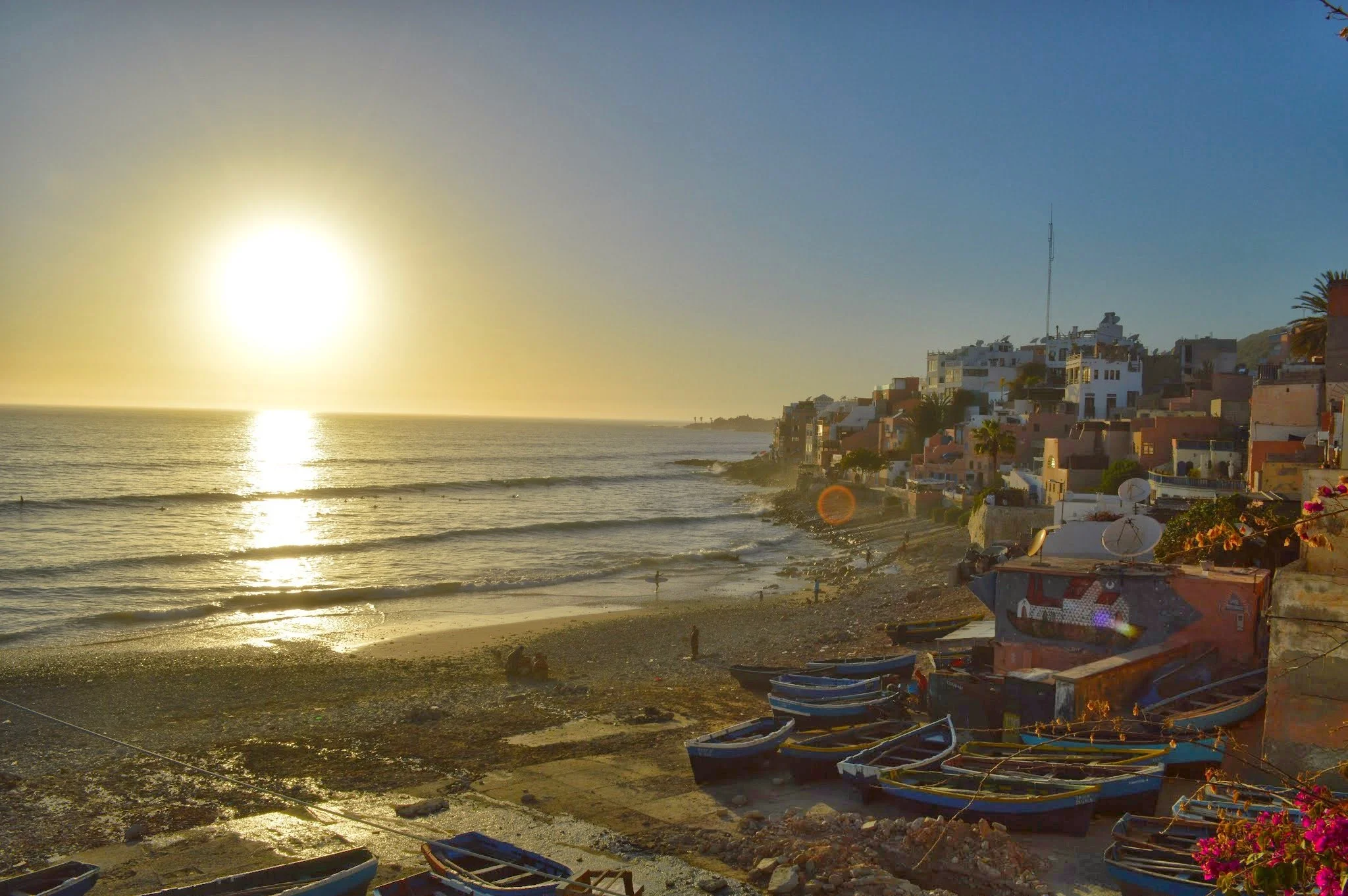 Sunset over a beach with boats in the foreground and a hillside with white residential buildings in the background.