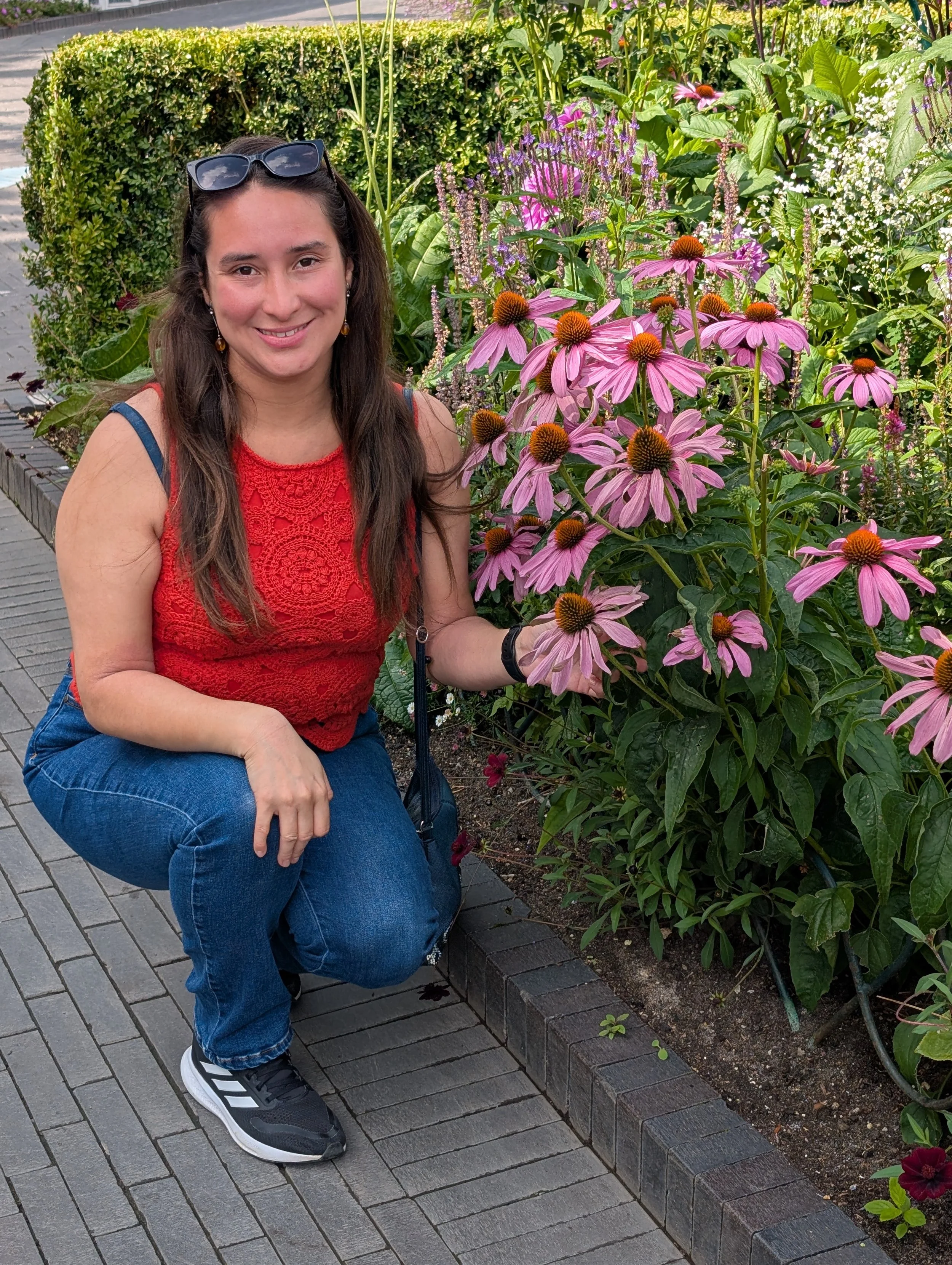 A smiling woman with long brown hair, wearing a red sleeveless lace top, blue jeans, and sneakers, kneeling on a brick sidewalk next to a flower bed filled with pink and purple flowers and green bushes.