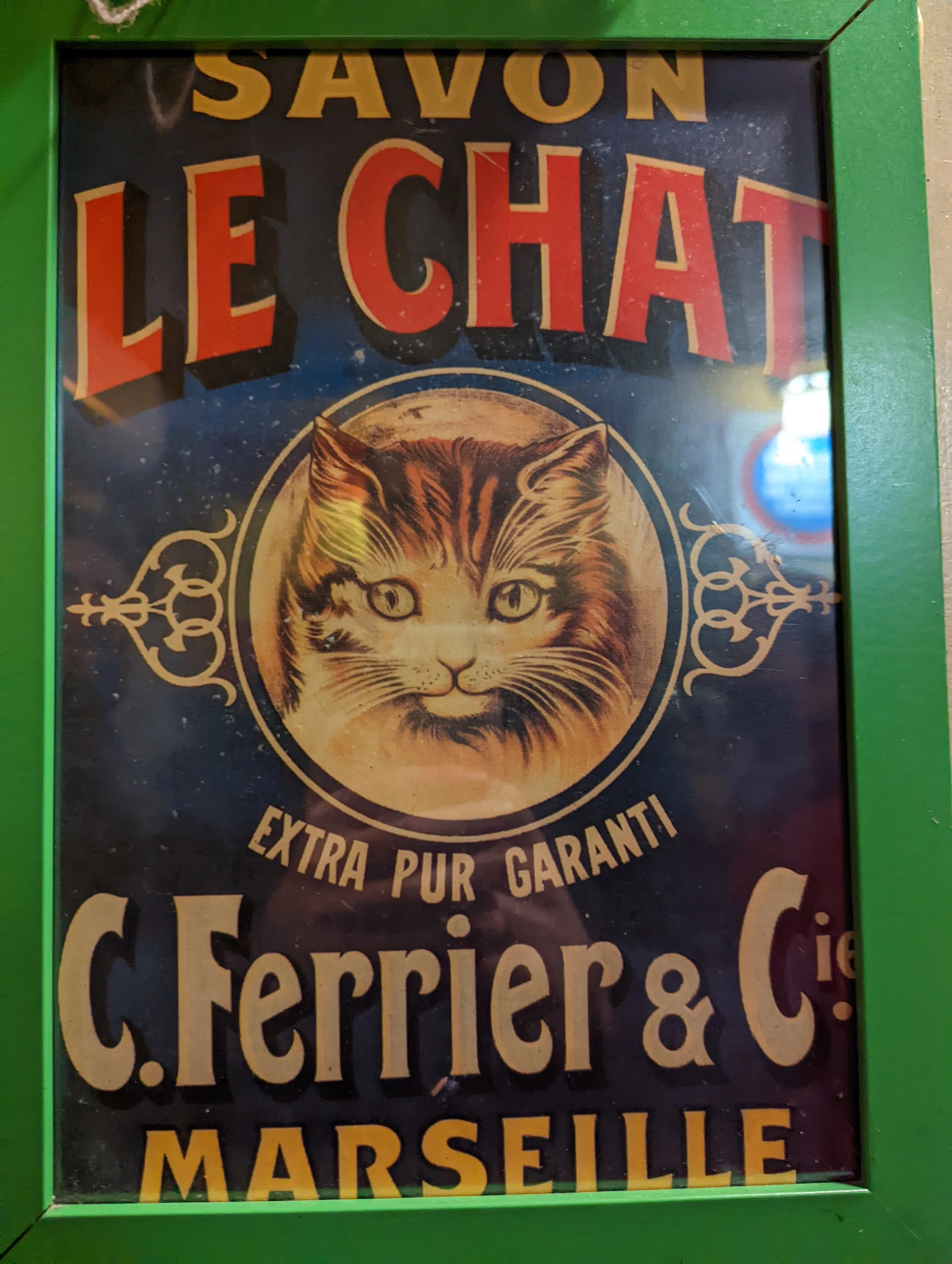 A framed sign advertising soap with a detailed illustration of a cat's face at the center, surrounded by ornate decorative elements, and text that reads: 'SAVON LE CHAT EXTRA PUR GARANTI' and 'marseille'.