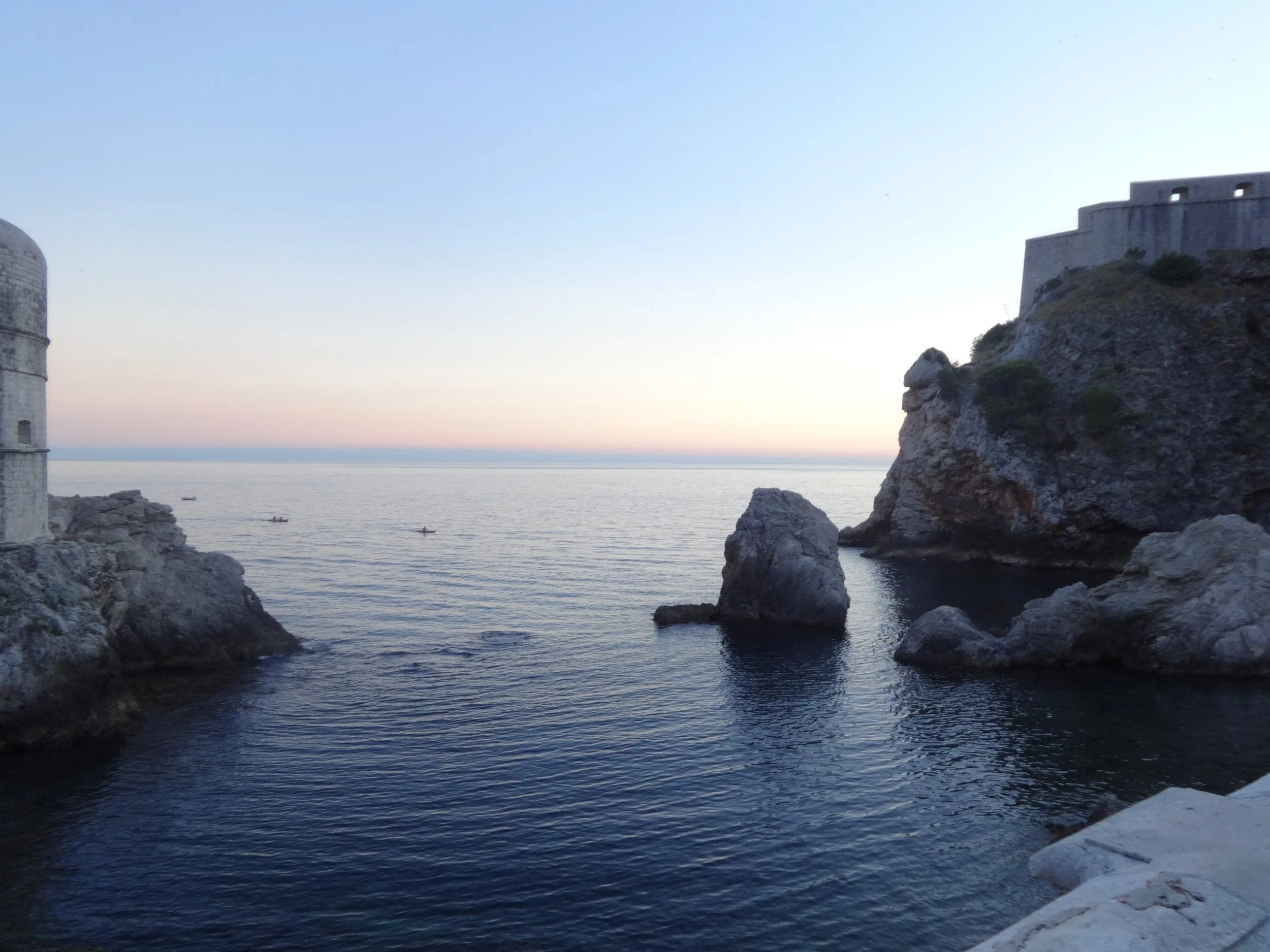 A coastal scene with rocky cliffs and a historic stone fortress overlooking the sea during sunset or dusk.
