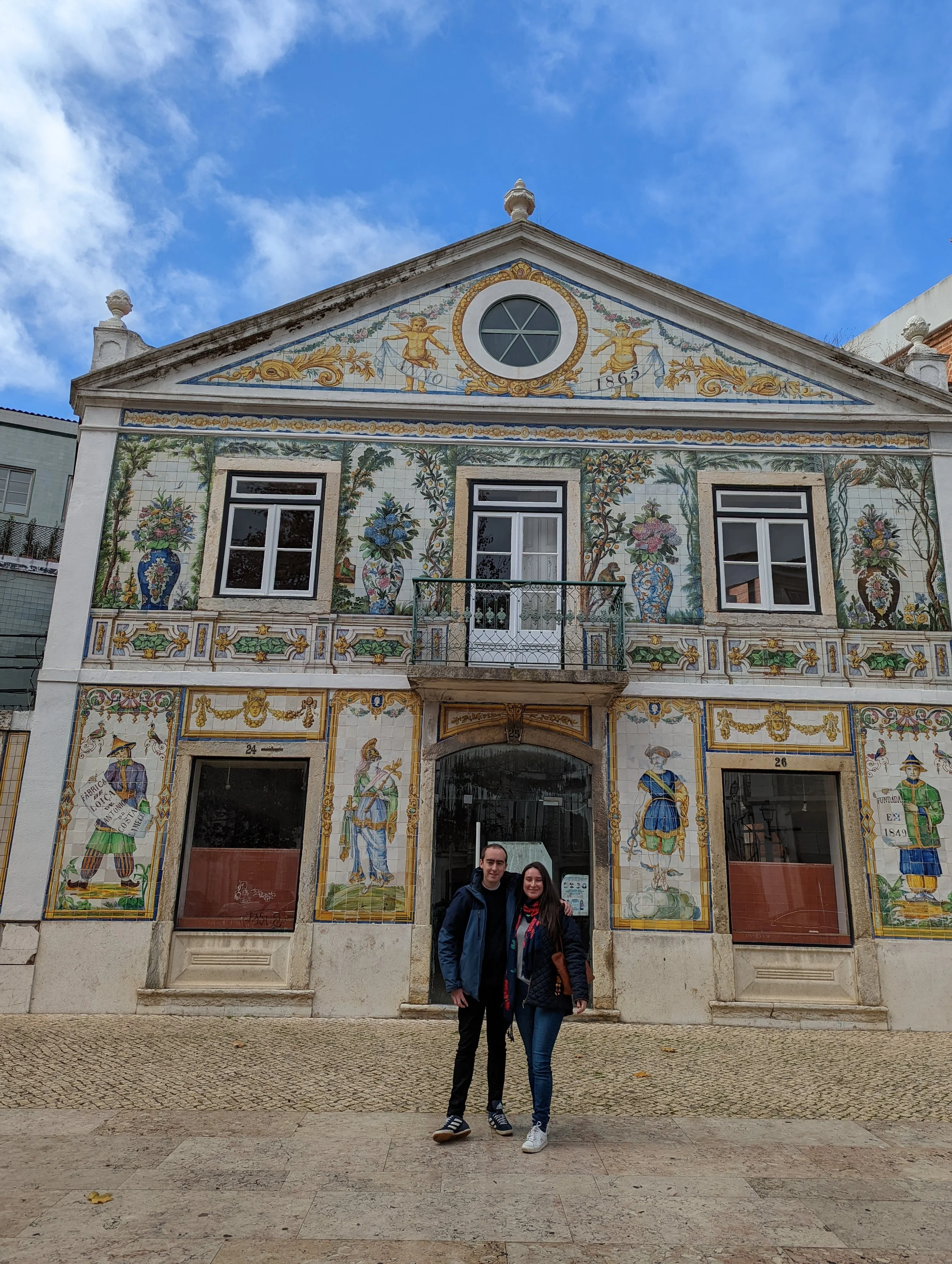 Picture of a historic building with a painted facade, featuring floral and decorative motifs, two people standing in front, overcast sky in the background, and cobblestone pavement.