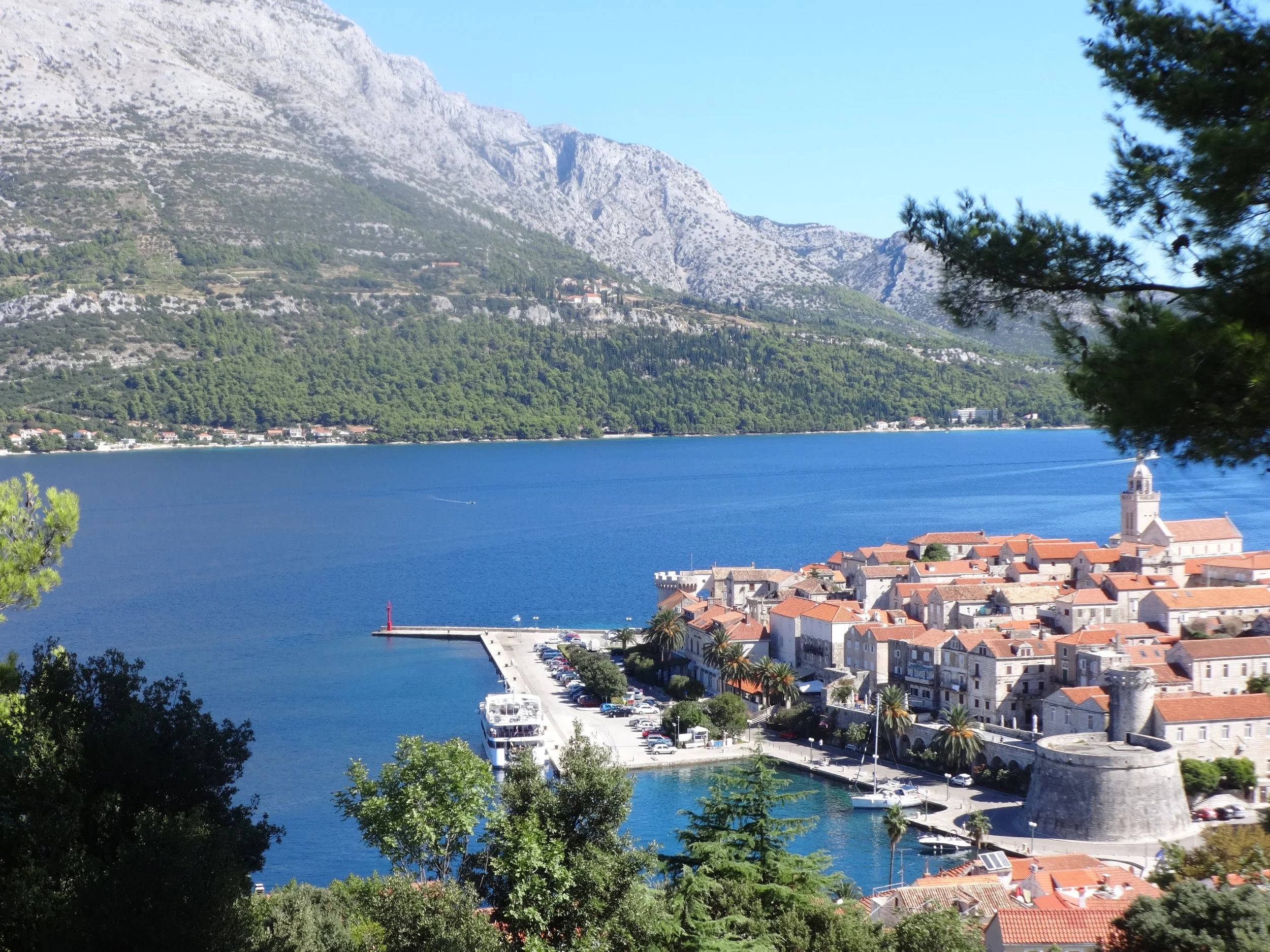A coastal town with red-tiled roofs along the water, a church steeple, and a mountain range in the background under a clear blue sky.