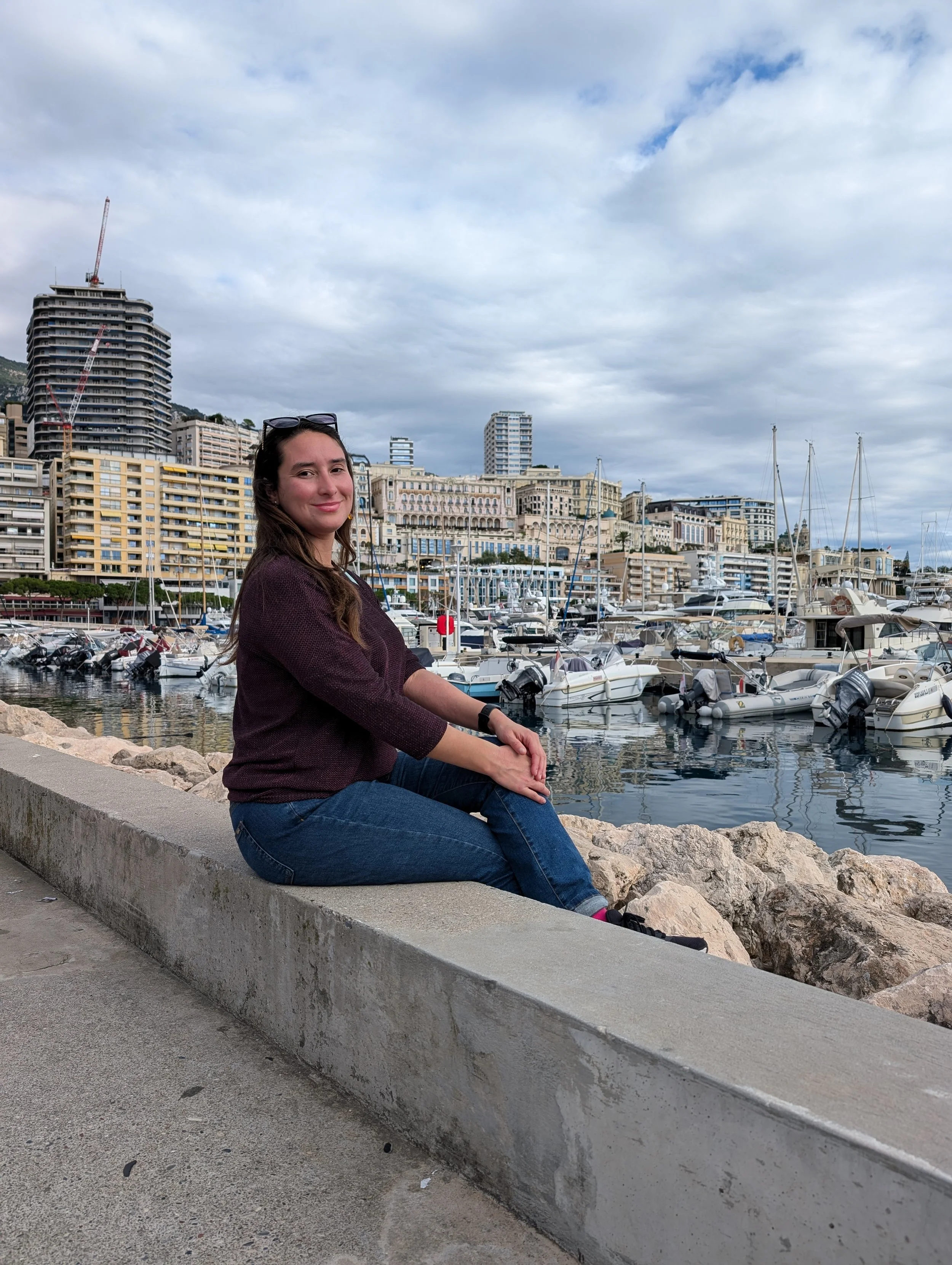 A young woman with long brown hair and sunglasses on her head sitting on a concrete barrier by a marina, with boats docked and tall buildings in the background.