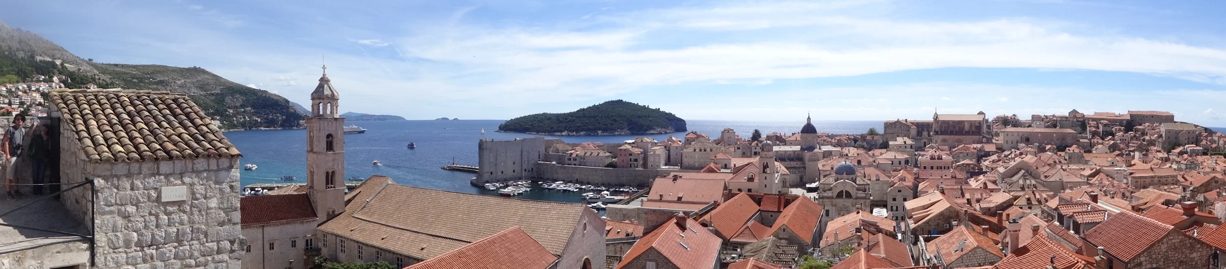 Panoramic view of a coastal city with stone buildings and orange rooftops, a harbor with boats, and an island in the background under a partly cloudy sky.