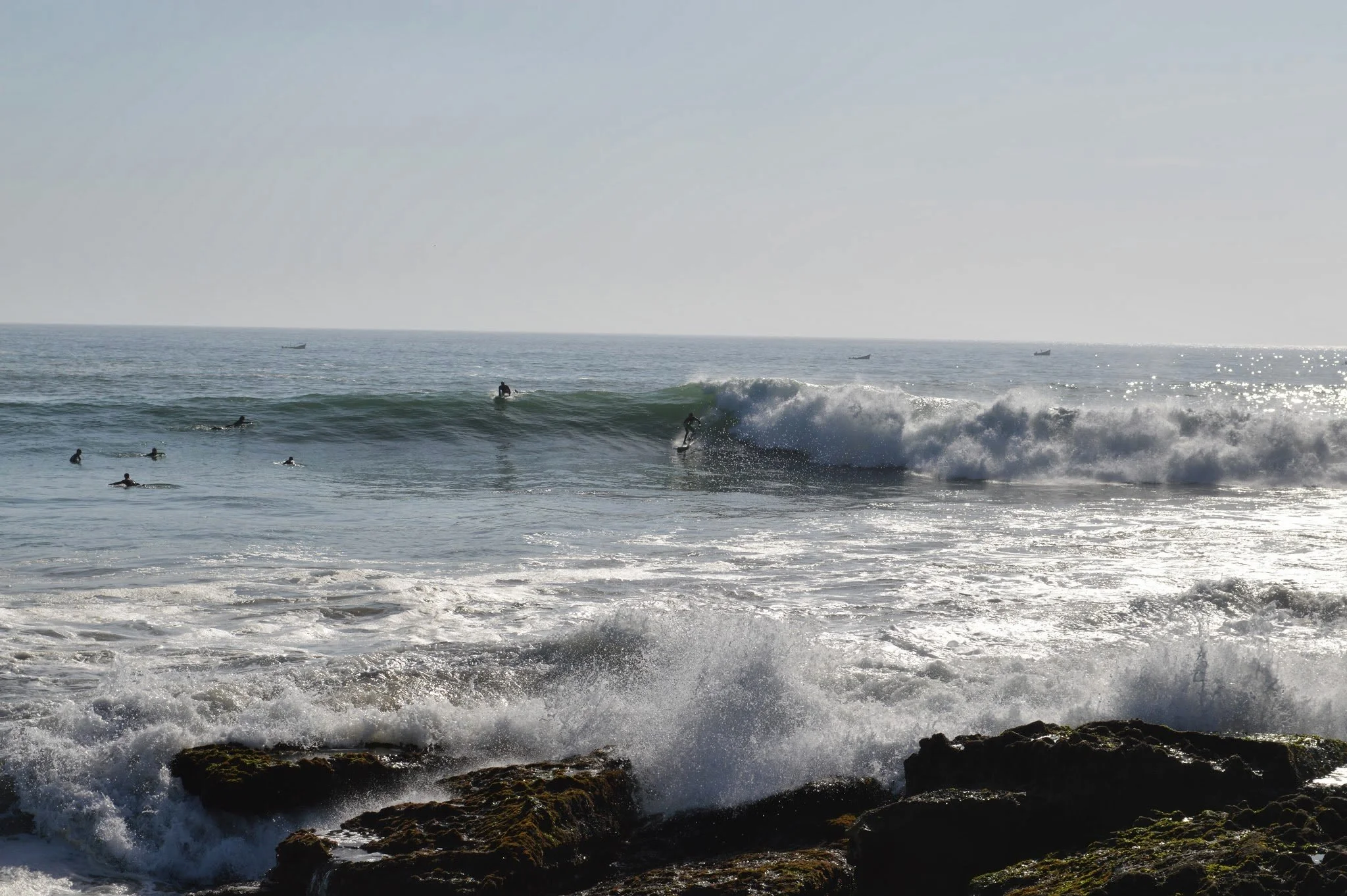 View of the ocean with waves breaking near the rocky shore, several surfers riding the waves, and a few boats on the horizon under a clear sky.