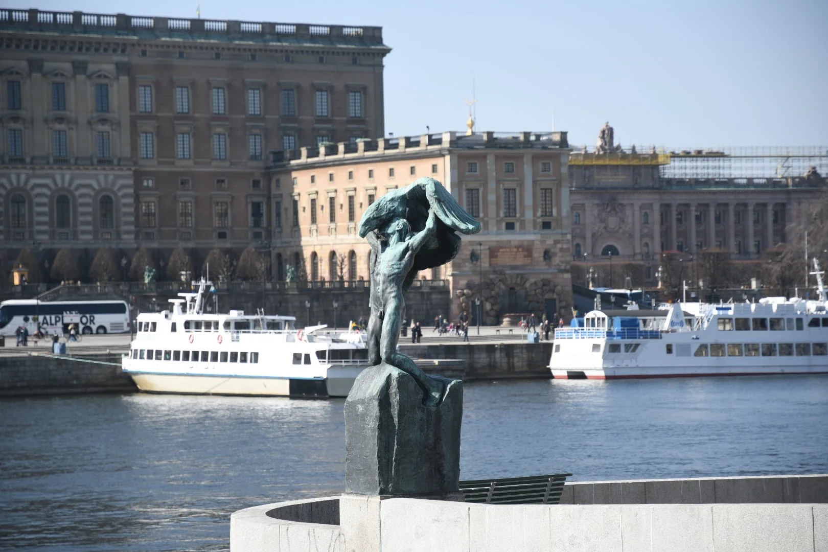 A bronze statue of a sailor holding a flag stands by a harbor with boats, in front of historic buildings across the water.