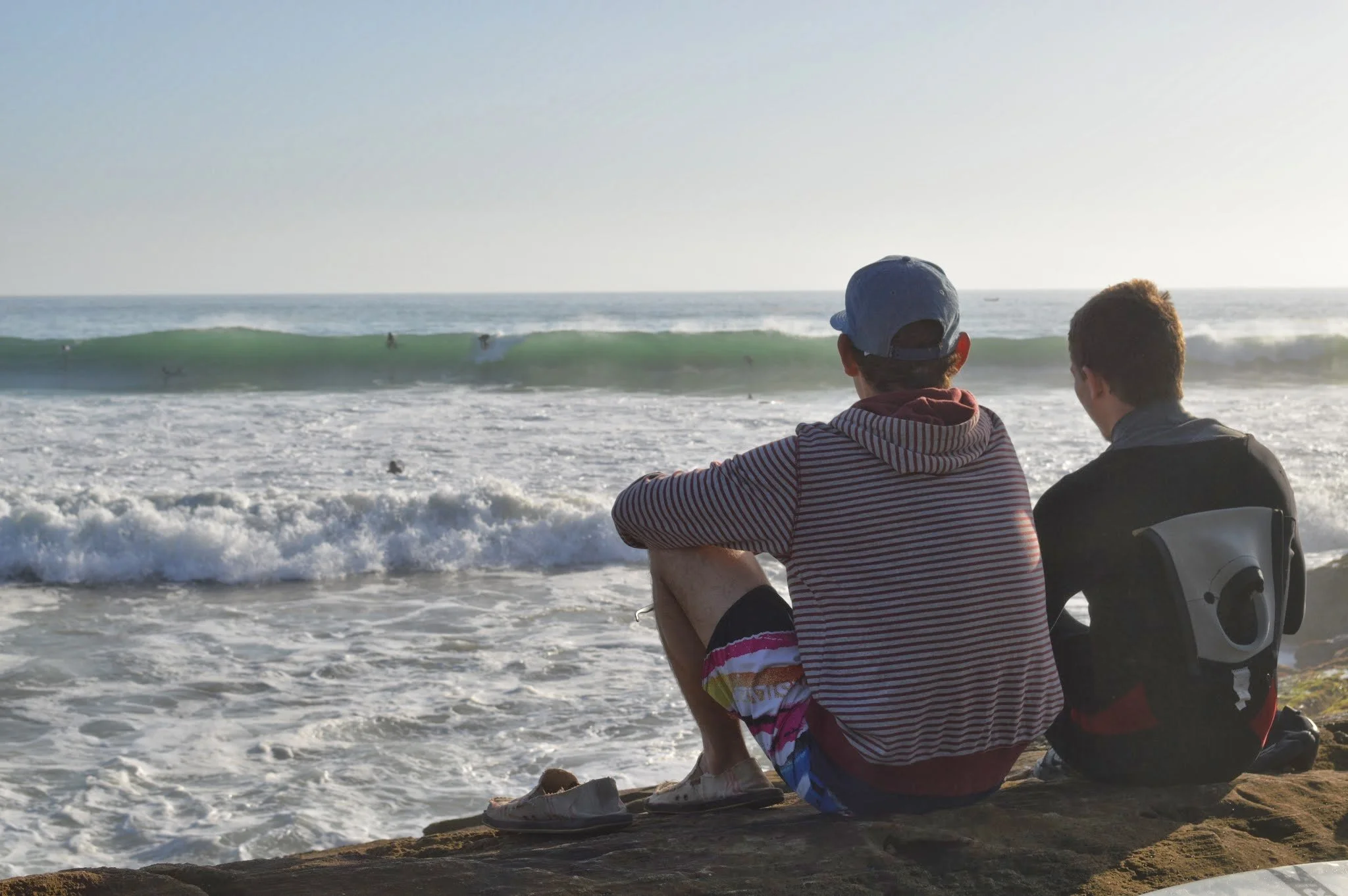 Two young men sitting on the beach, facing the ocean, watching surfers ride waves