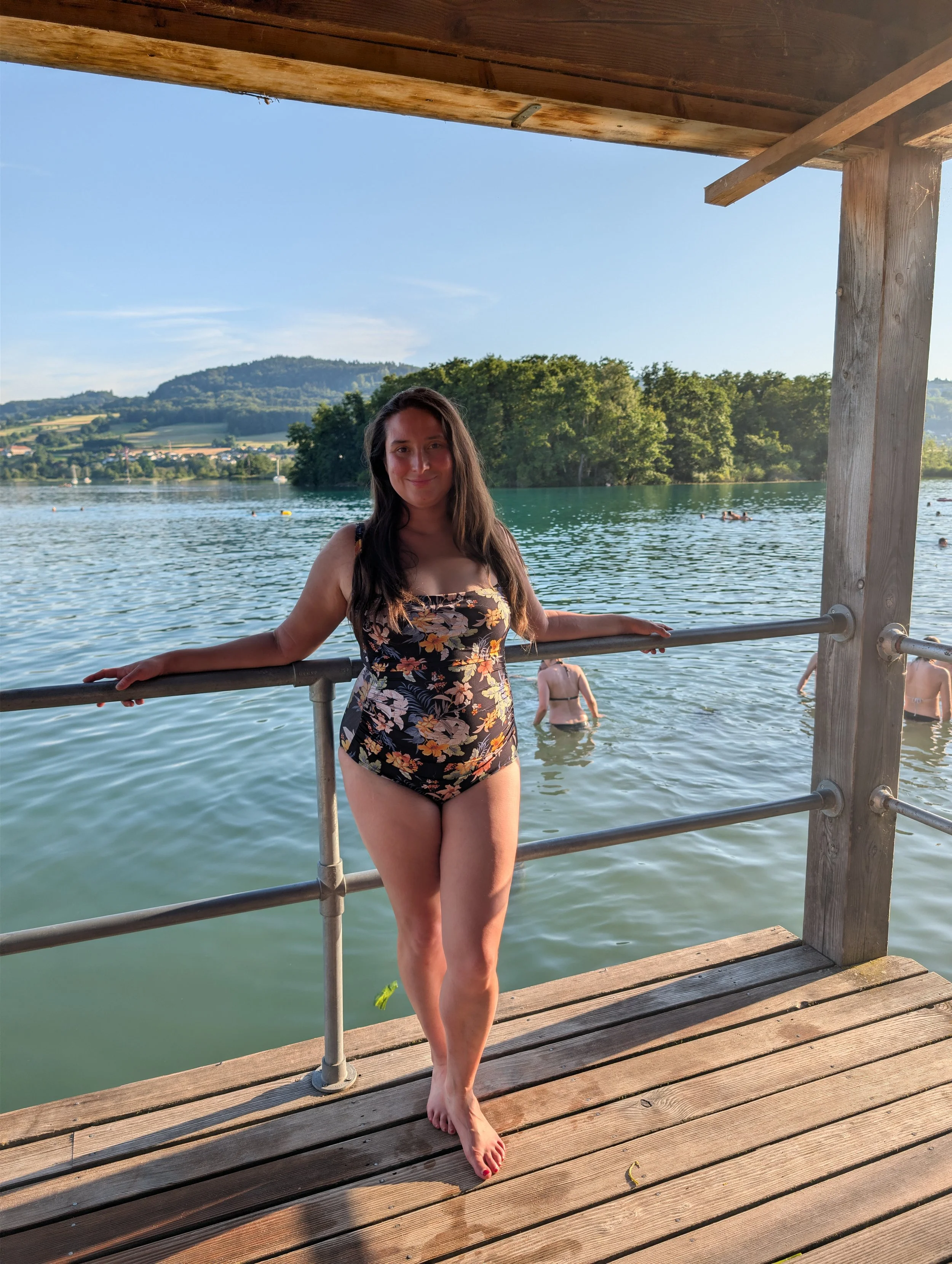 A woman in a floral swimsuit standing on a wooden dock by the water, with trees, hills, and a few people swimming in the background.
