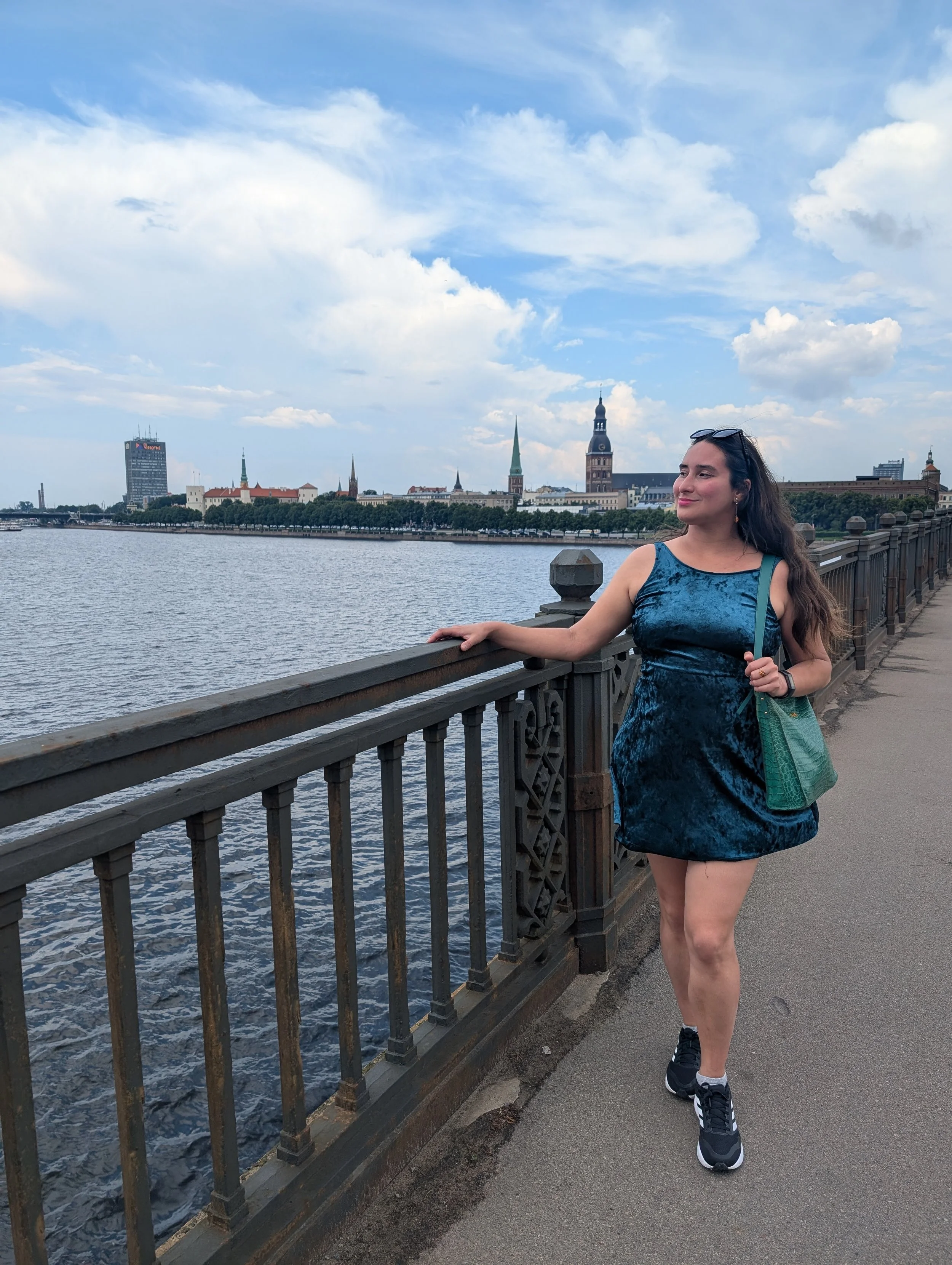 A woman in a blue dress and black sneakers walking along a riverside promenade with a metal railing. In the background, a cityscape with historic buildings, church steeples, and a modern hotel under a partly cloudy sky.