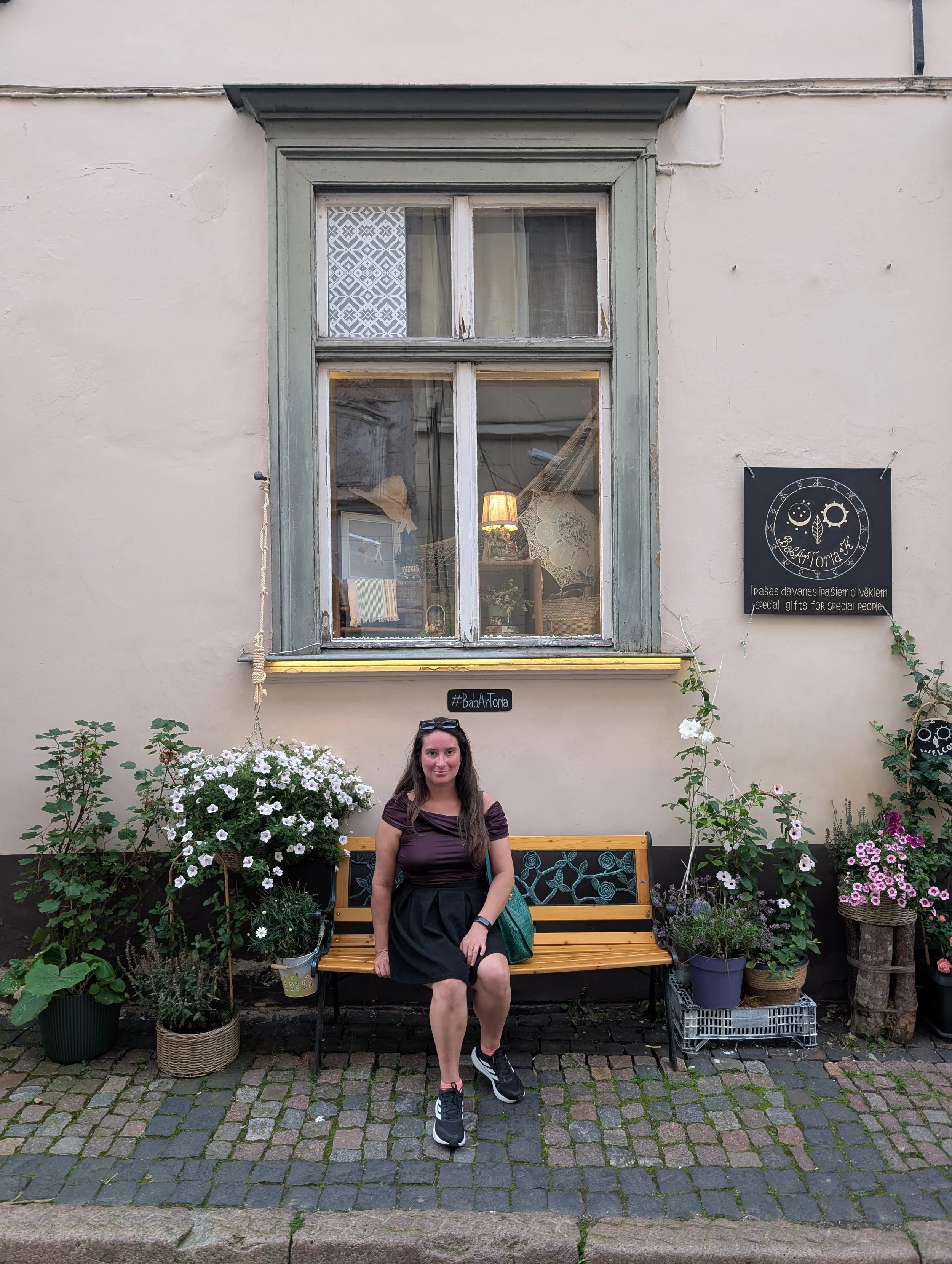 A woman sitting on a wooden bench in front of a building wall decorated with potted plants and flowers, with a window above her and a sign that reads 'BīskapiArToria'.