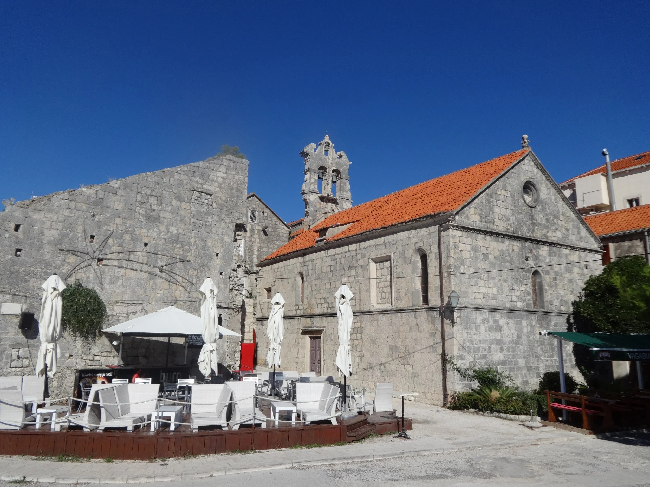 Stone church with red tile roof and a bell tower under a clear blue sky, with outdoor seating and umbrellas in the foreground.