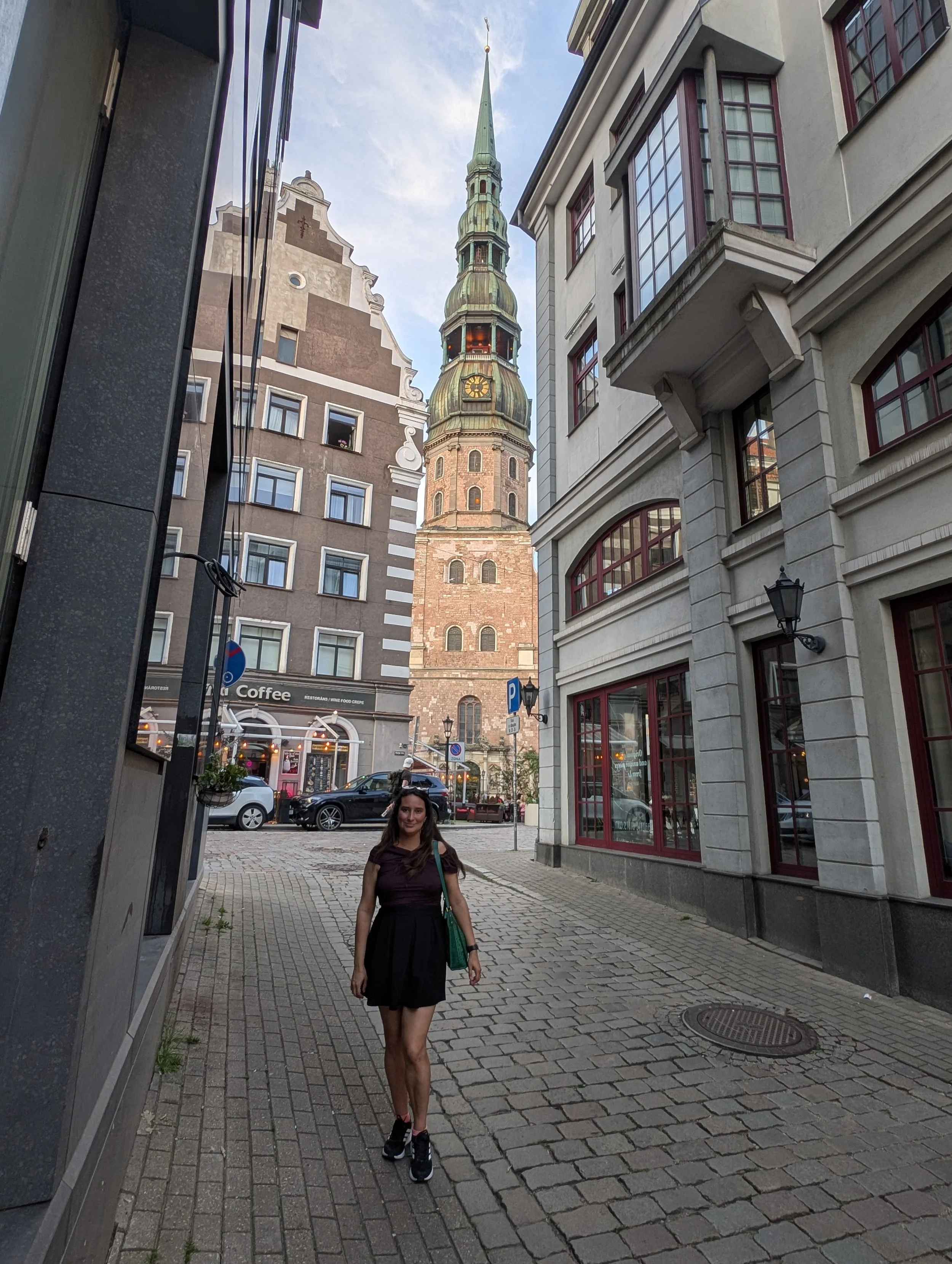A woman with long brown hair, wearing a black dress and black sneakers, walking on a cobblestone street in an urban area with historic buildings and church tower in the background.