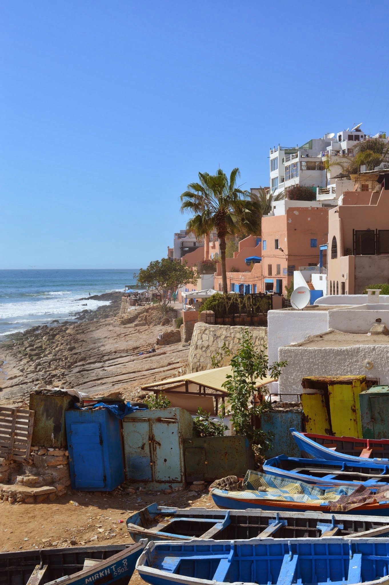 Colorful buildings on a hillside above a rocky shore with boats and debris in the foreground, and a blue sky in the background.