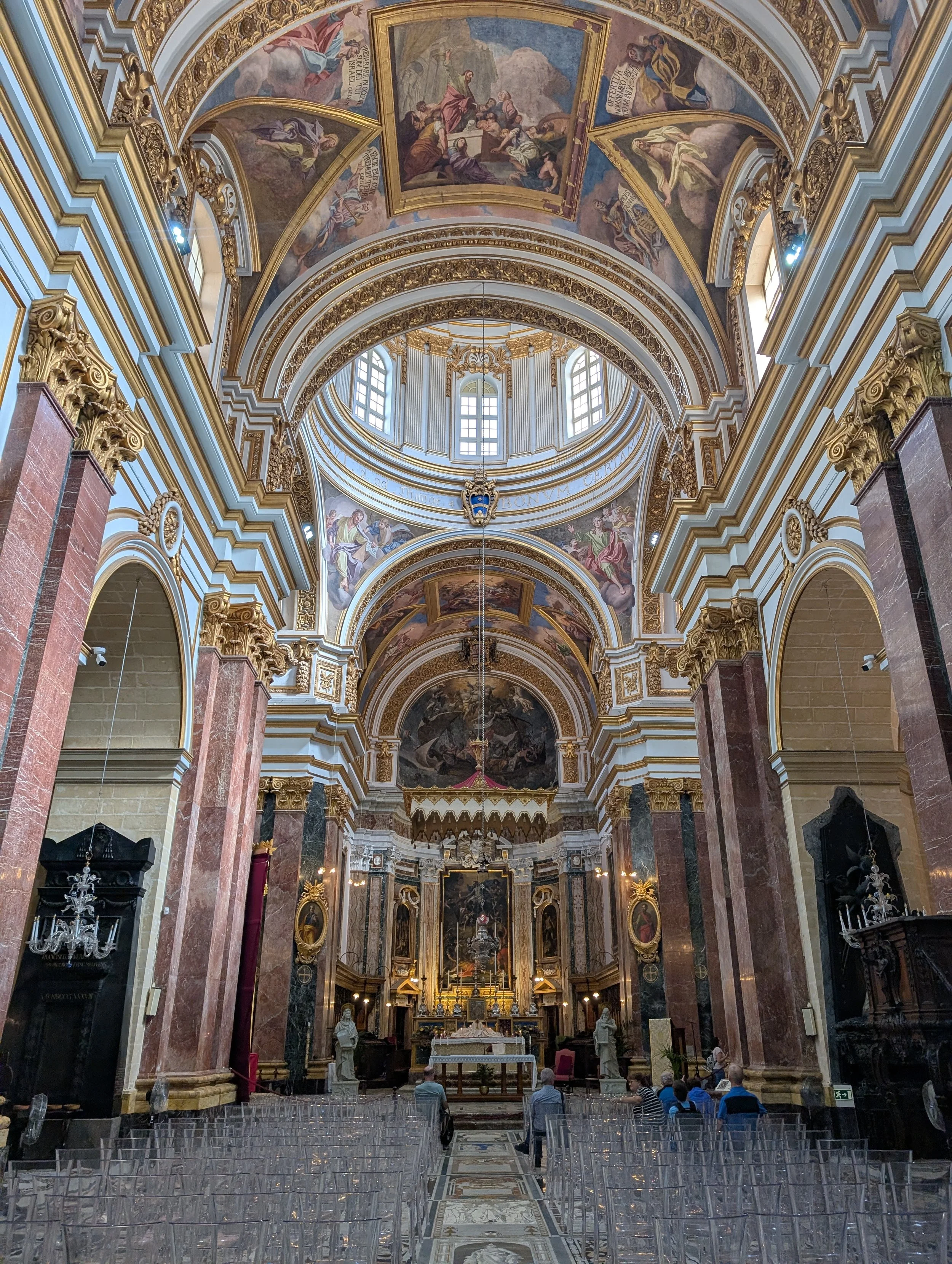 Interior of a grand church with ornate gold and white decoration, arched ceiling with painted religious scenes, marble columns, and an altar at the front with statues and paintings. People are seated facing the altar.