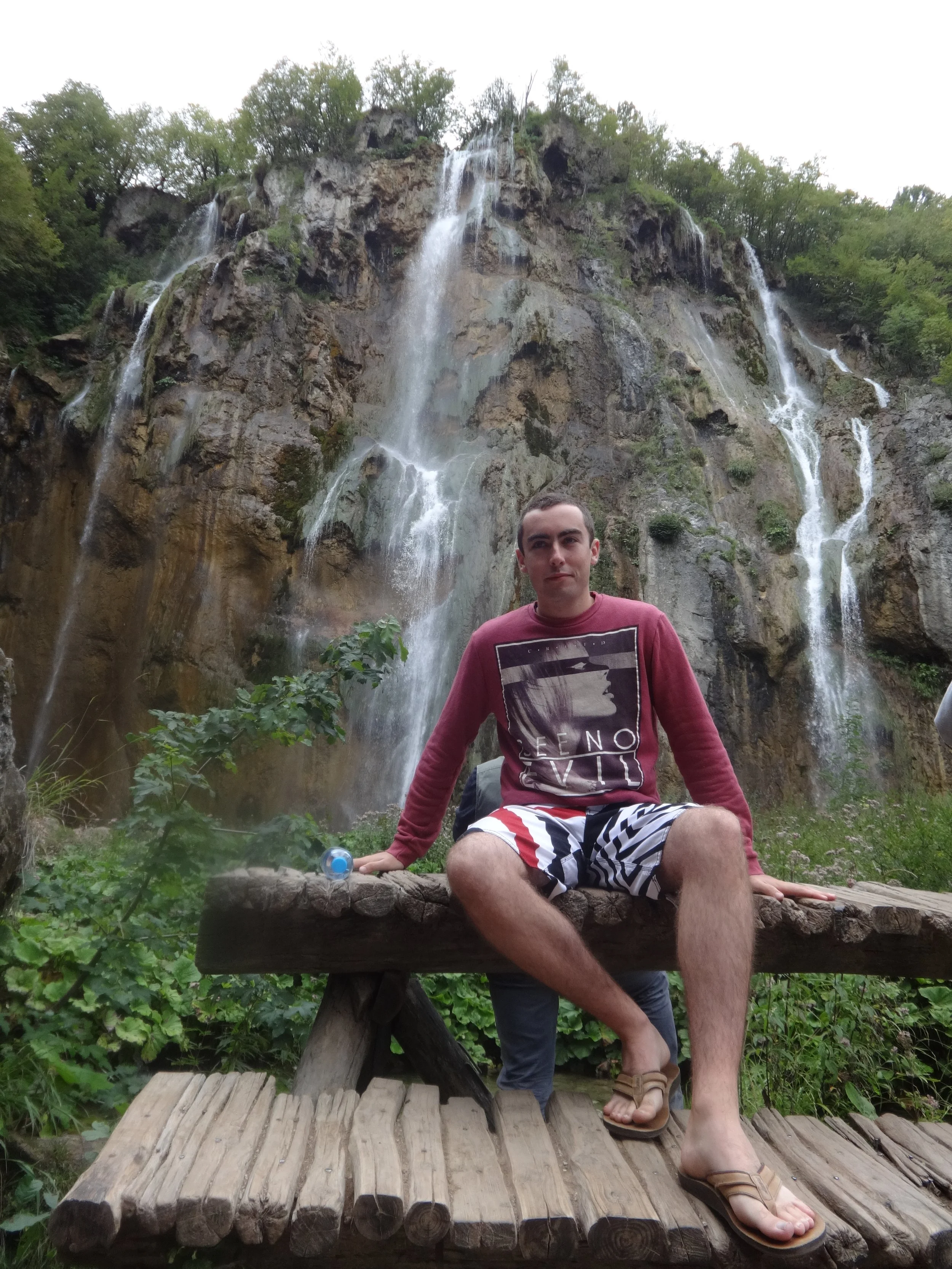 A young man sitting on a wooden bridge in front of a waterfall, wearing a maroon shirt and striped shorts, with lush greenery surrounding him.