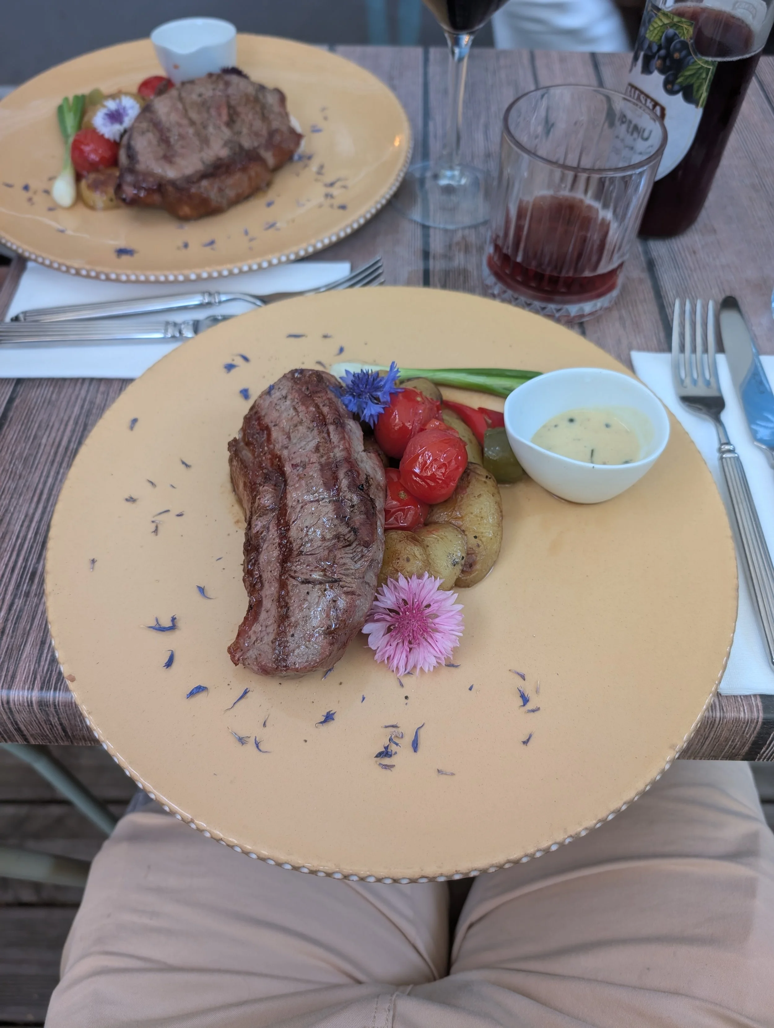 A top view of a person sitting at a wooden table with two plates of steak, roasted vegetables, a small dish of sauce, and flowers. There are also a glass of red wine and a bottle of wine on the table.