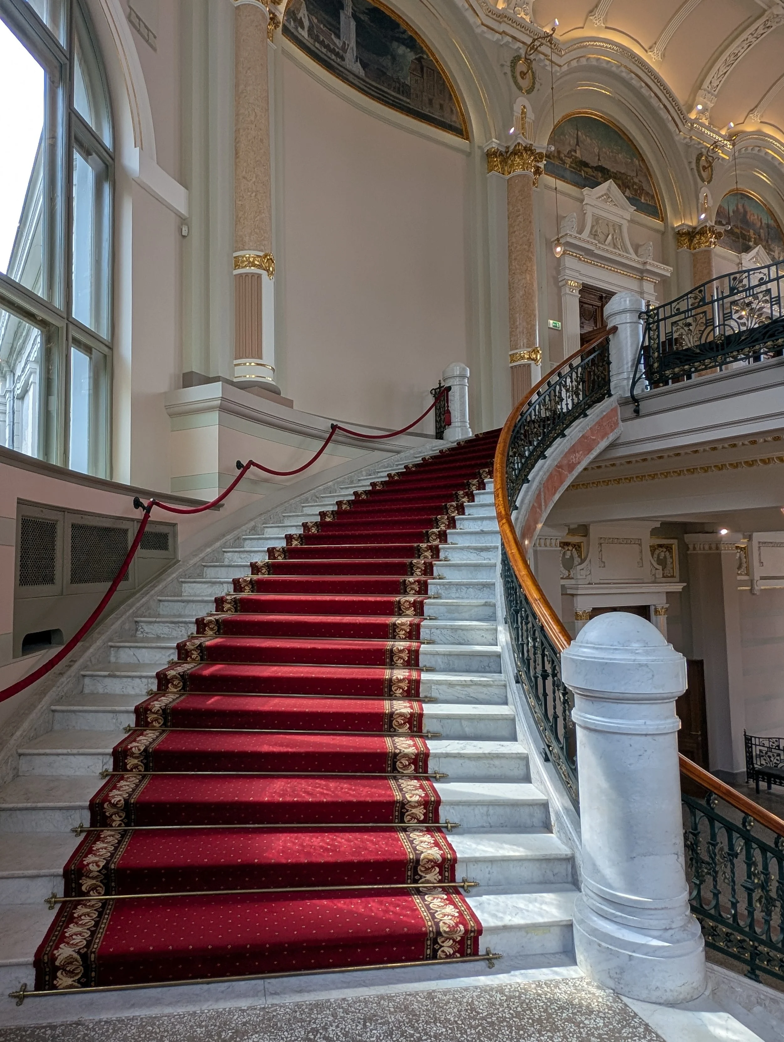 Interior of a grand staircase with marble steps covered by a red carpet runner with gold and floral borders, brass rods, and a curved wrought iron and wood handrail, leading to an upper floor in a lavishly decorated building with ornate white and gol