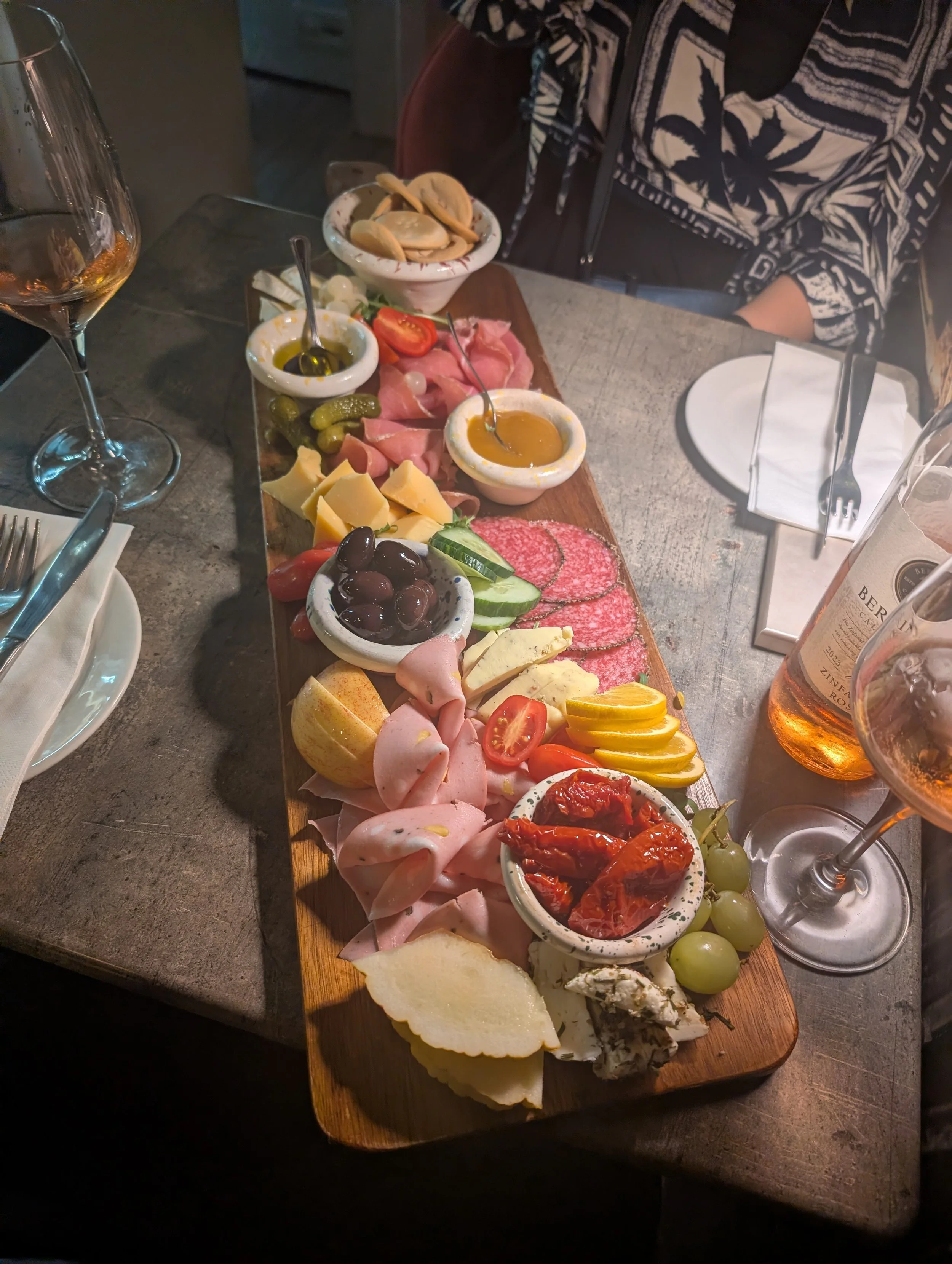 Charcuterie board with assorted cheeses, deli meats, tomatoes, olives, pickles, sliced vegetables, and bread at a dining table with wine glasses.